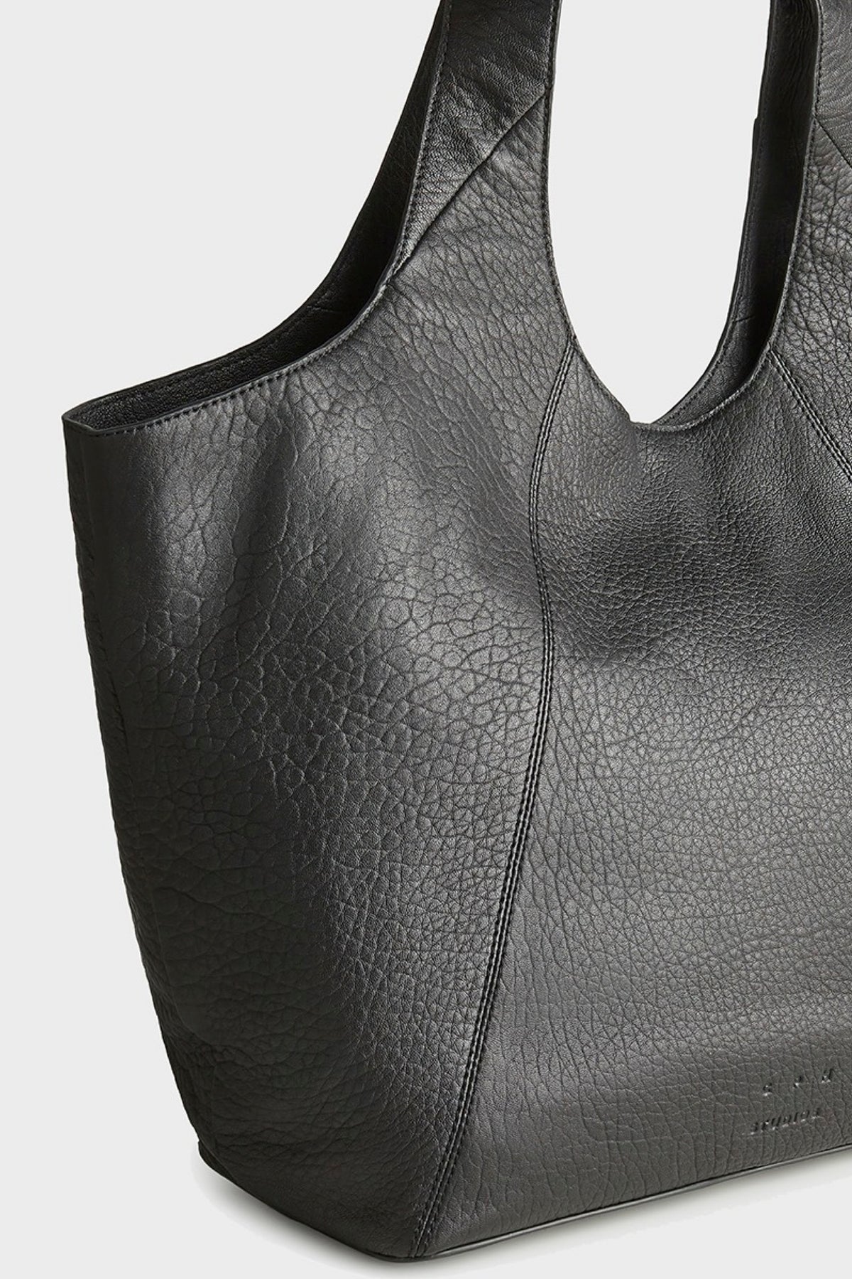 A close-up of a black pebbled leather tote bag with double shoulder strap, shown against a light gray background.