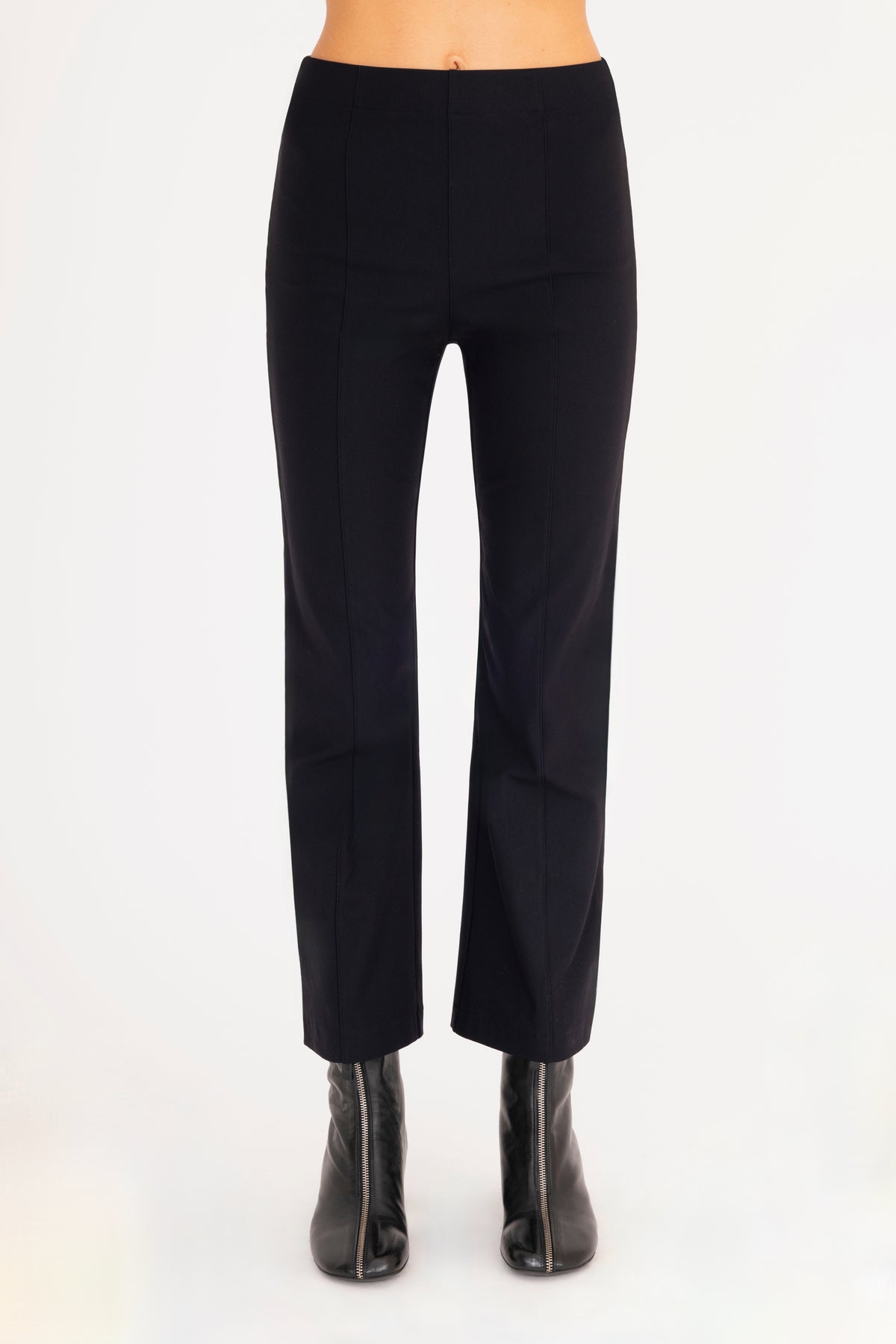 A front view of a model wearing  black flared pants with black vegan leather contrast piping and black leather ankle boots, set against a plain white background.