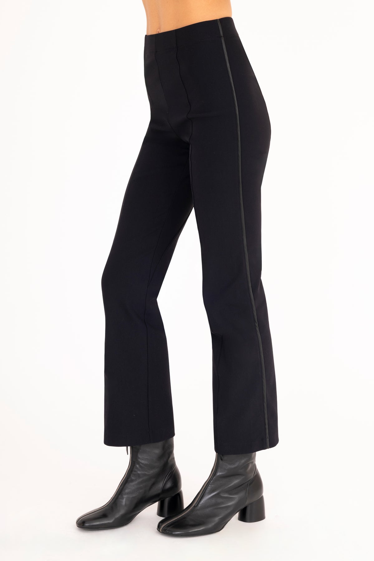 A side view of a model wearing  black flared pants with black vegan leather contrast piping and black leather ankle boots, set against a plain white background.