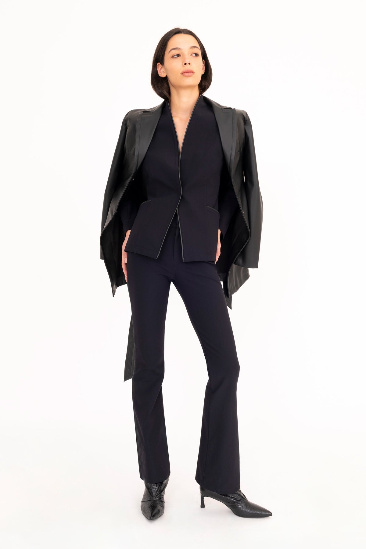 A model stands against a solid white studio background wearing a black tailored suit consisting of a structured blazer and boot-leg pants, with a black vegan leather trench coat draped over her shoulders and black pointed-toe heels.