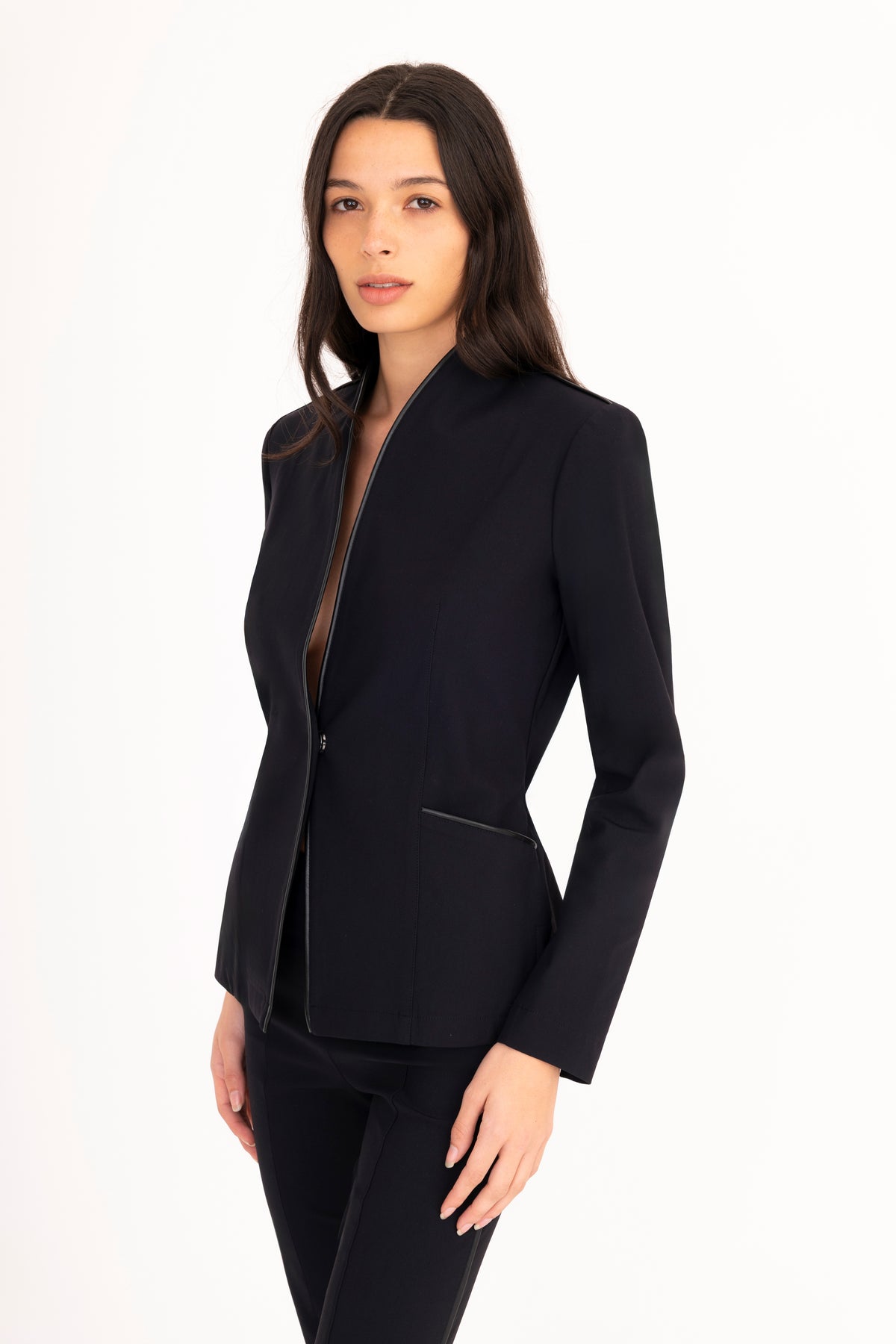 A woman modeling a structured black blazer with a stand collar and a single front button, paired with black trousers, against a plain white studio background.
