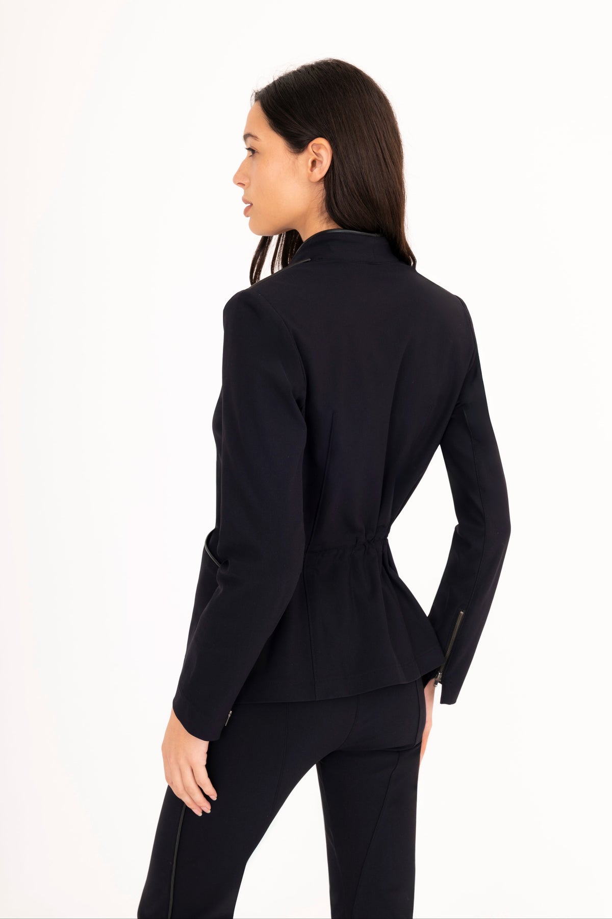 A woman shown from the back wearing a tailored black blazer with a structured collar and peplum waist, paired with black trousers, against a plain white studio background.