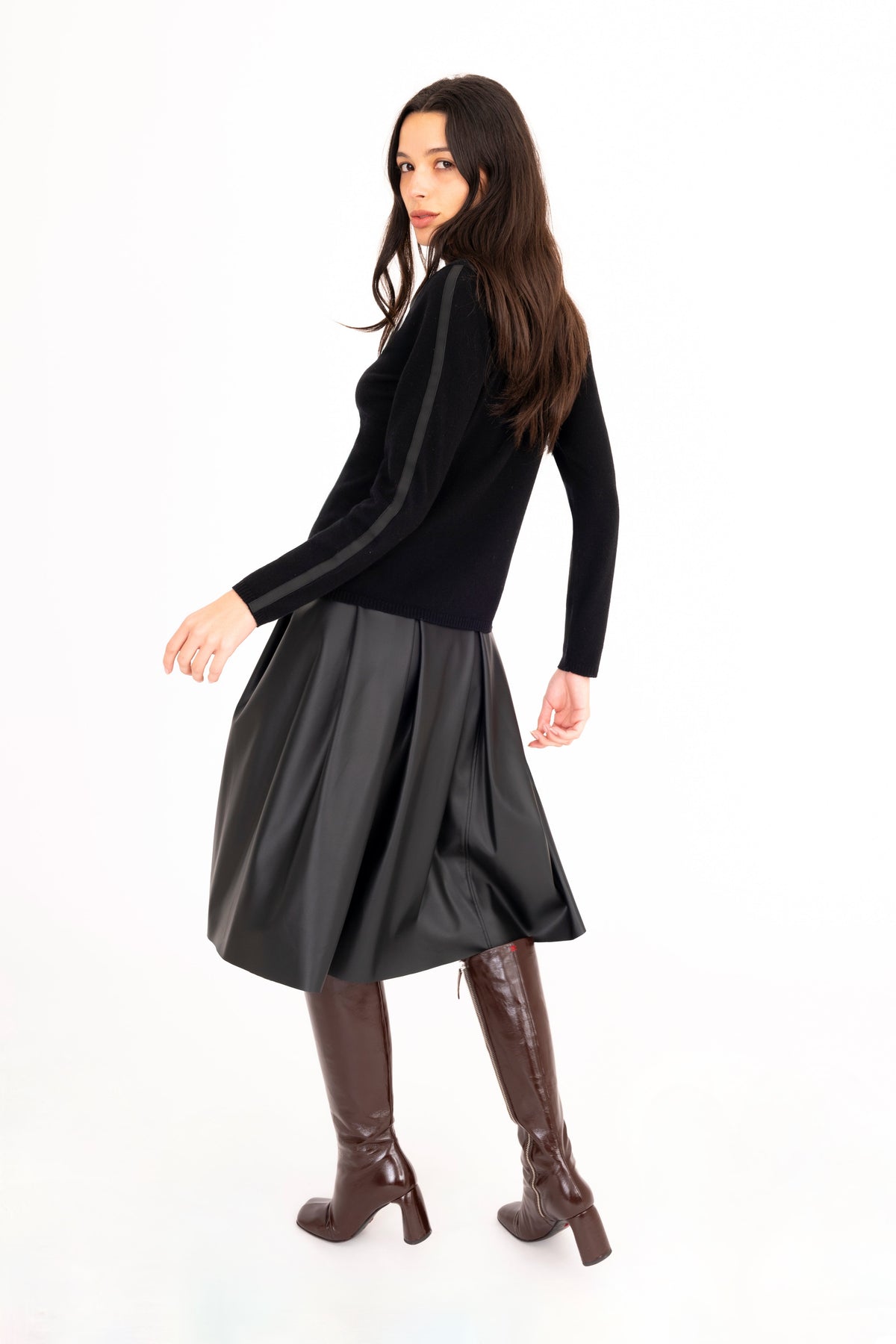A woman modeling a black long-sleeve cashmere turtleneck sweater, a black vegan leather pleated midi skirt, and dark brown knee-high boots against a plain white studio background.