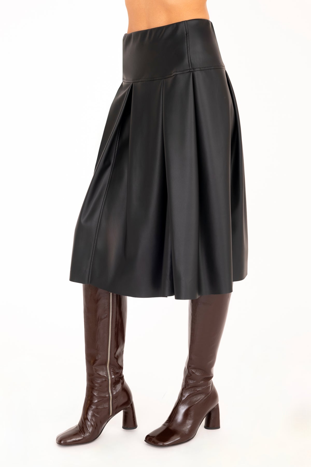 A black vegan leather pleated A-line skirt shown on a model from the waist down, paired with dark brown knee-high leather boots against a plain white background.
