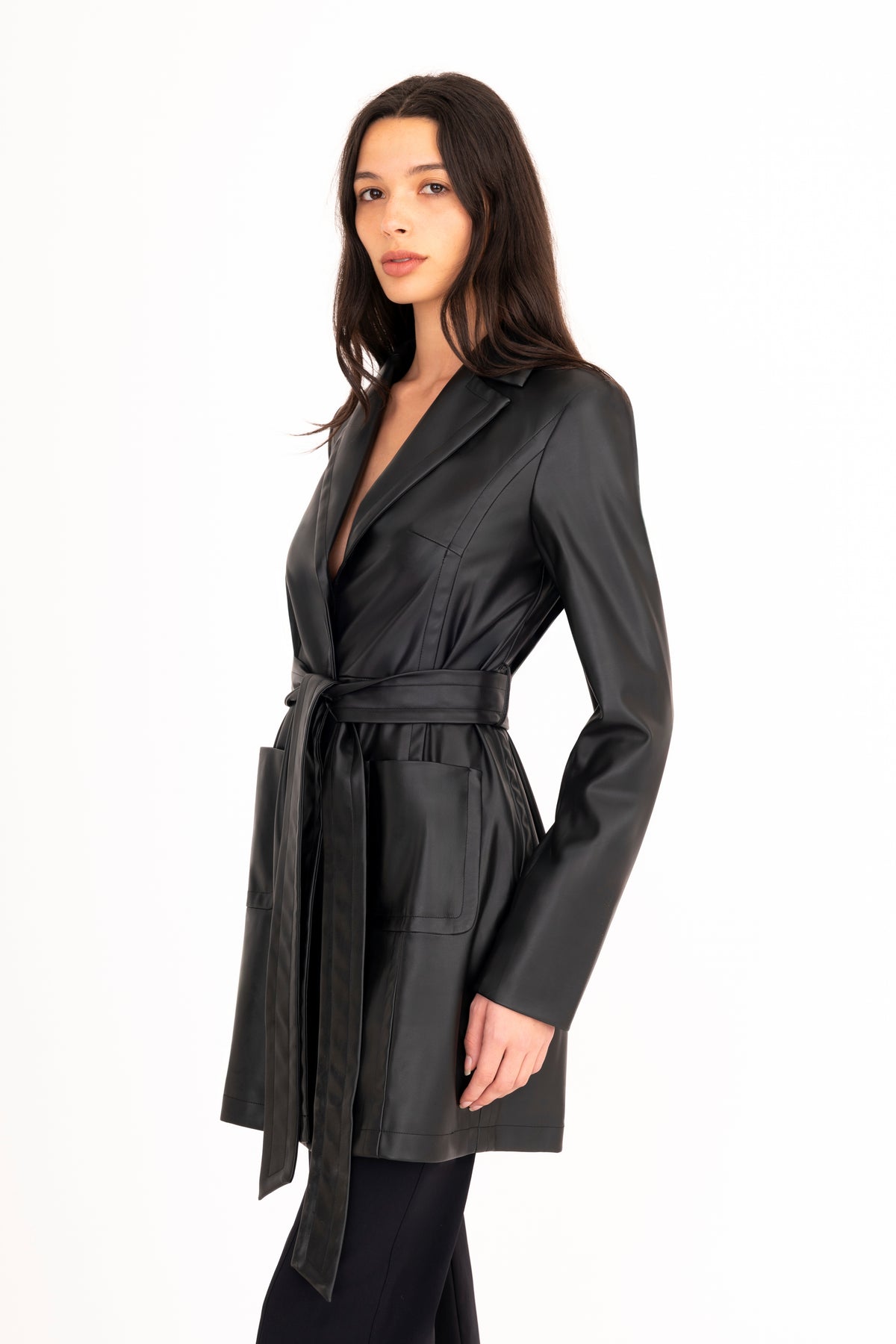 A woman modeling a black vegan leather trench coat with a collar, waist tie, and front pockets, paired with matching black pants, against a plain white studio background.