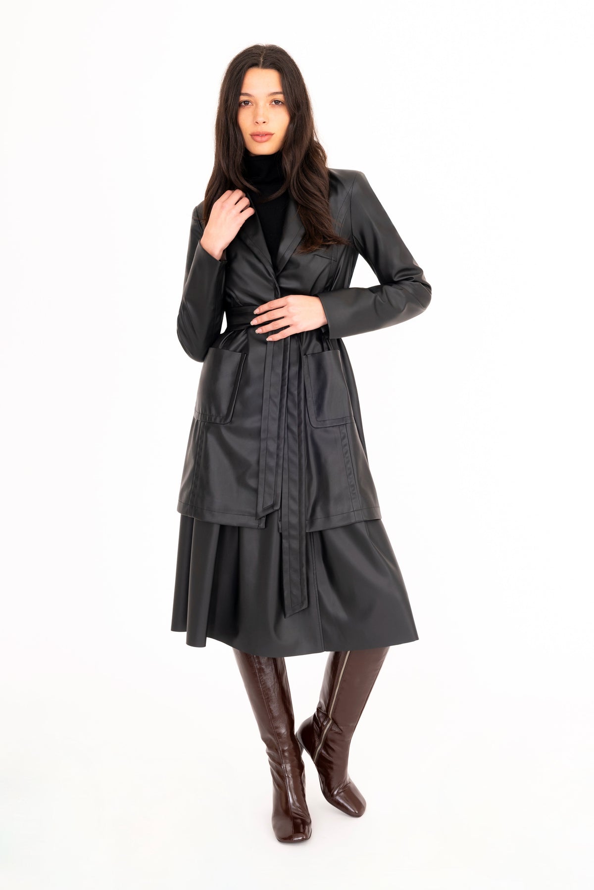 A woman modeling a black vegan leather trench coat and a pleated skirt, paired with brown knee-high heeled boots, against a plain white studio background.