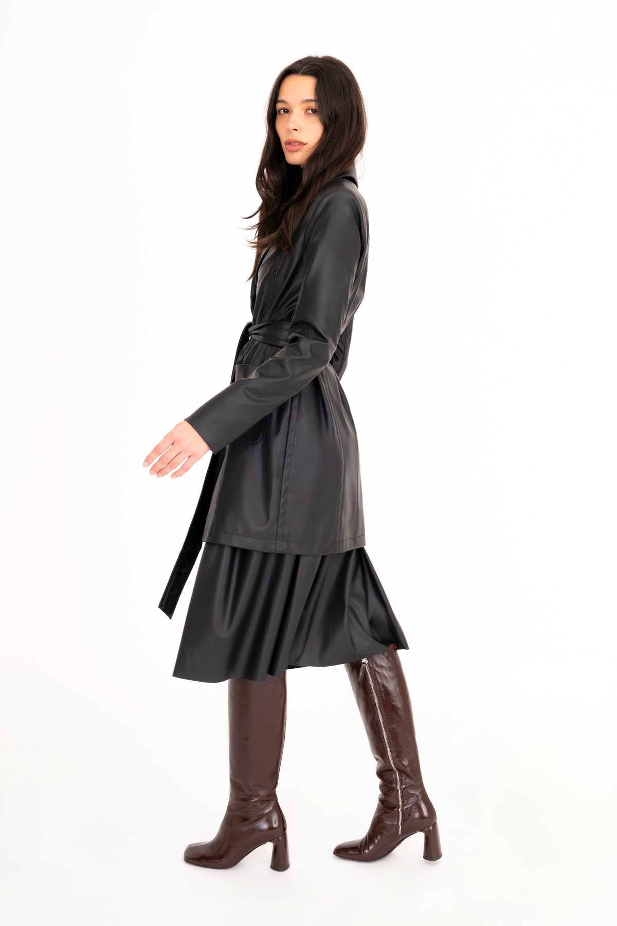 A woman modeling a black vegan leather trench coat and a pleated skirt, paired with brown knee-high heeled boots, against a plain white studio background.