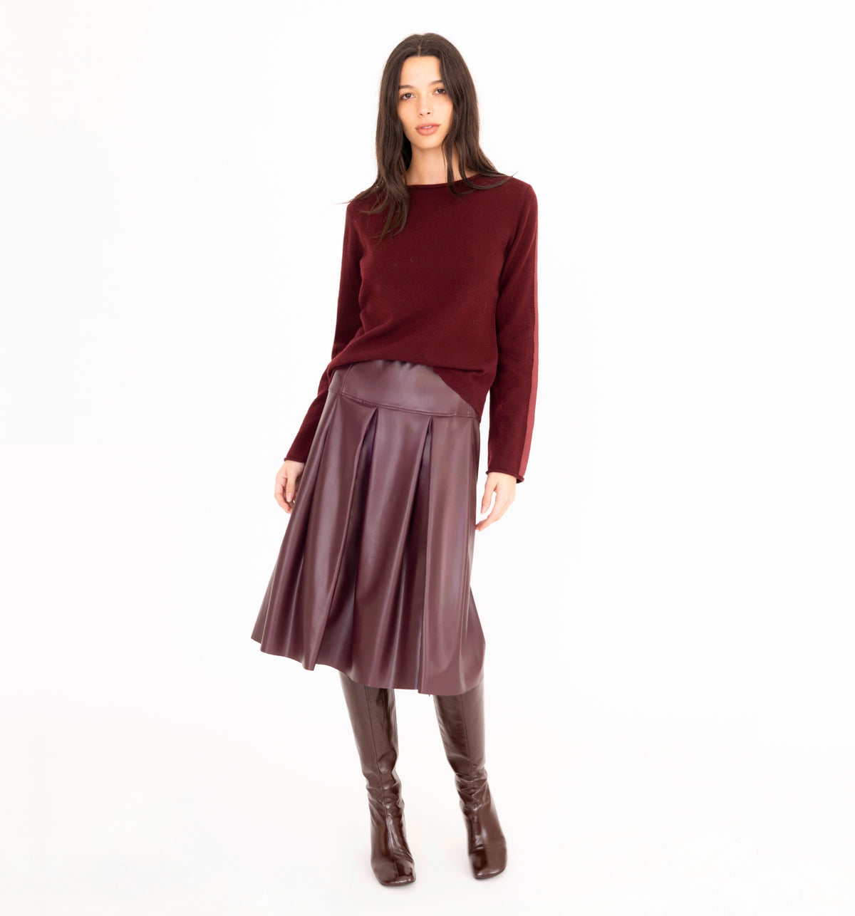 A model stands against a plain white studio background wearing a long-sleeve burgundy cashmere sweater, a pleated burgundy vegan leather midi skirt, and dark brown knee-high boots.