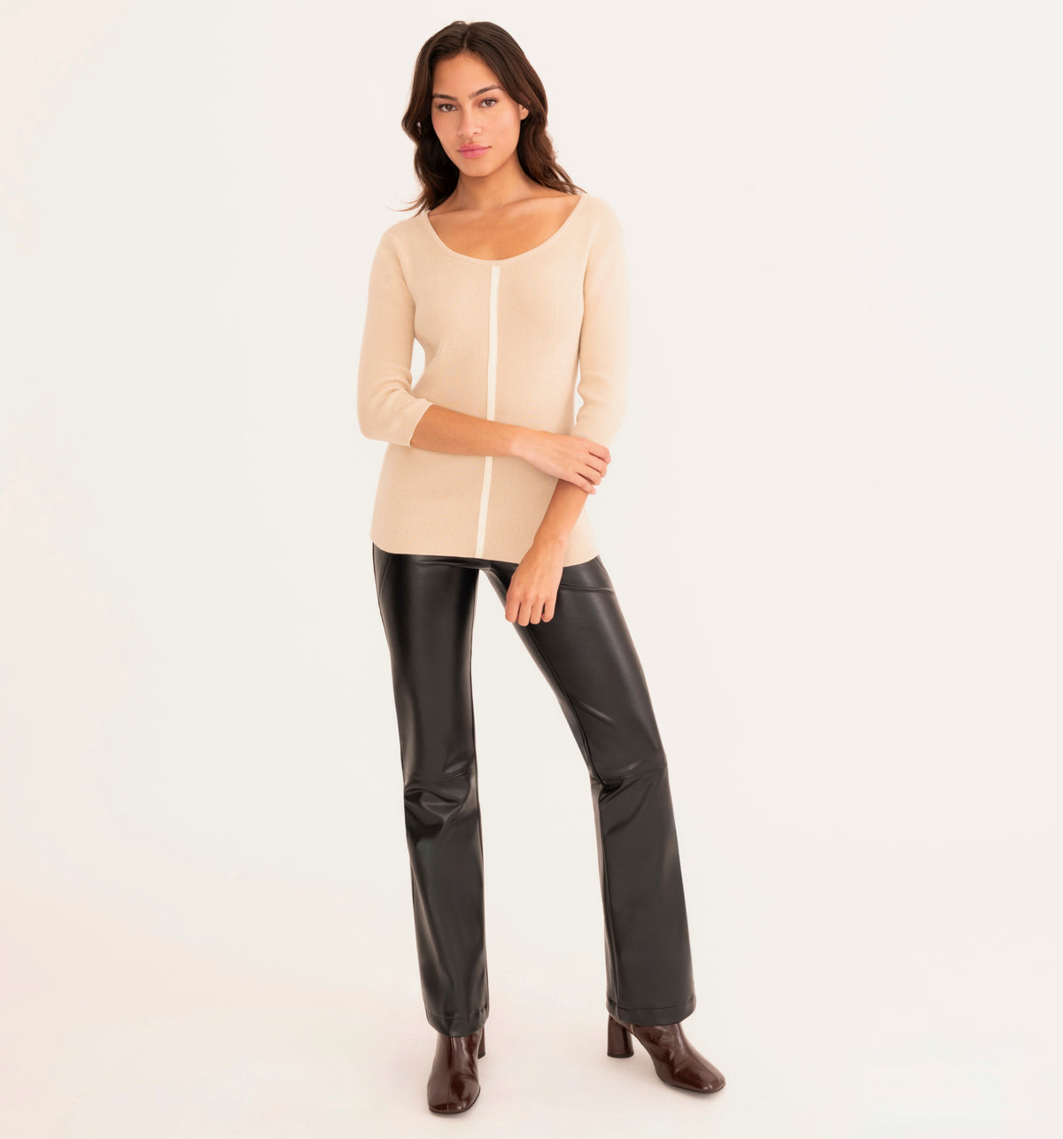 A woman modeling a beige knit top with a vertical white stripe down the center, paired with black faux-leather pants and brown ankle boots, against a plain white studio background.