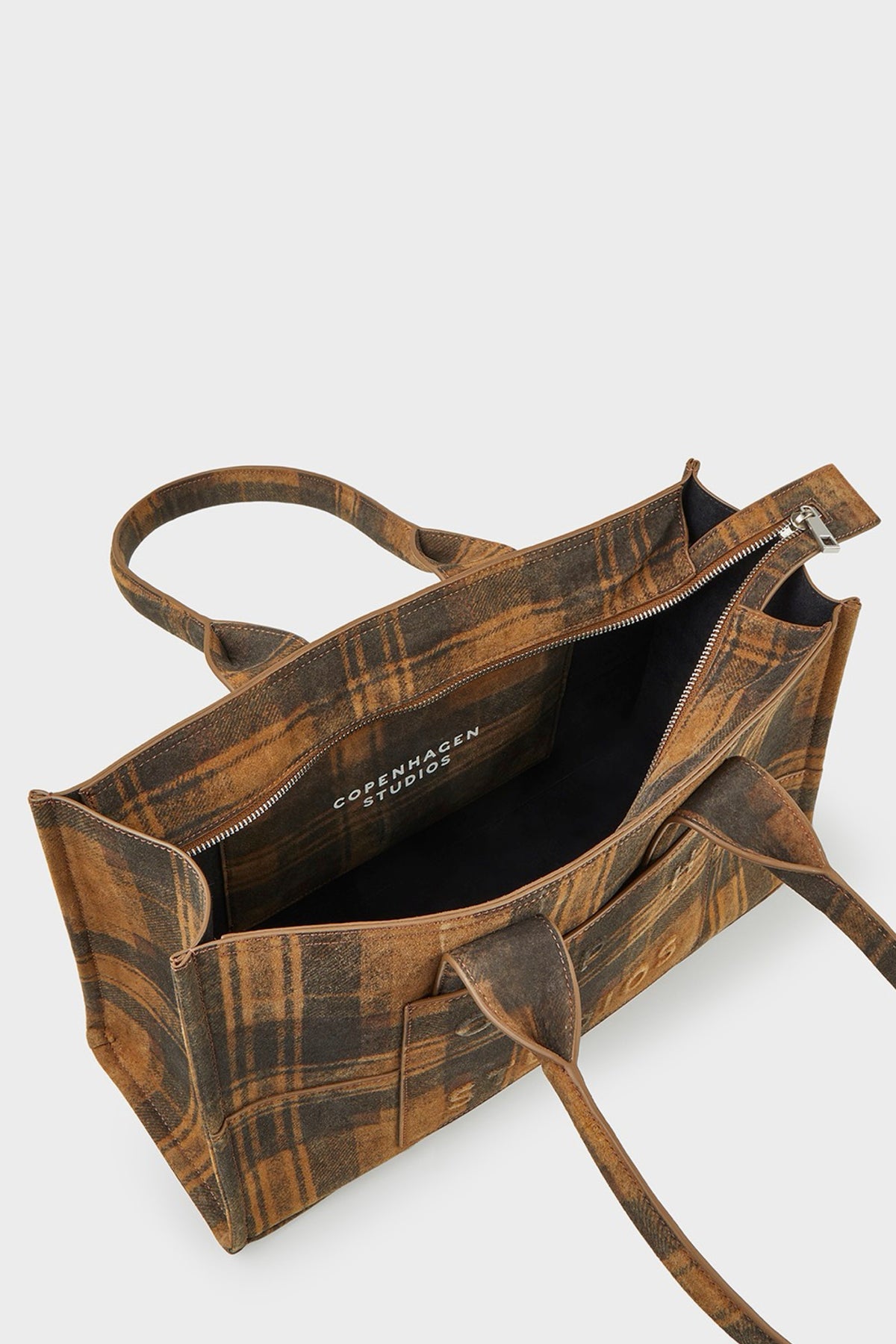 A brown and tan plaid suede tote bag shown open from a top-down angle, revealing a black interior lining with the brand name 'COPENHAGEN STUDIOS' printed inside. The bag features two top handles and a zippered closure, set against a clean, light gray background.