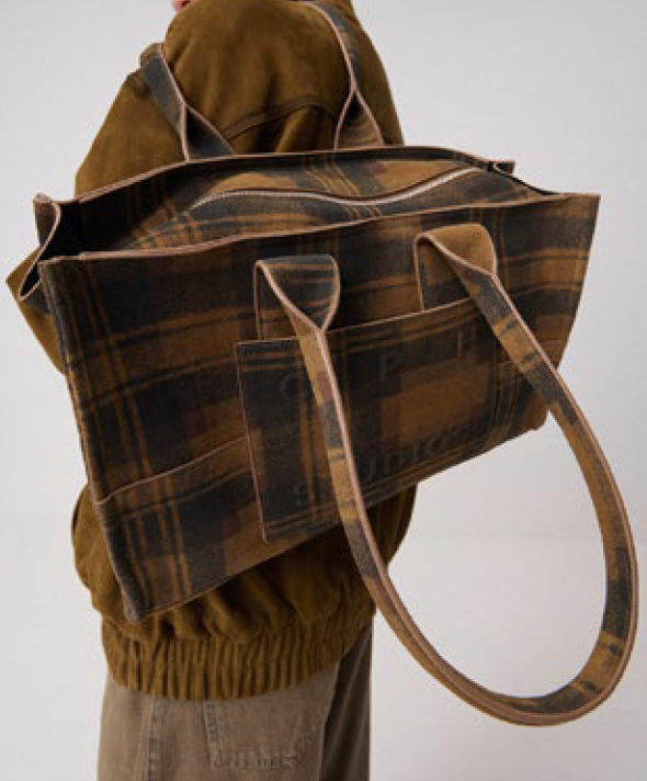 A large brown and tan plaid suede tote bag with matching leather handles, shown being worn over the shoulder of a person wearing a brown jacket against a plain light background.