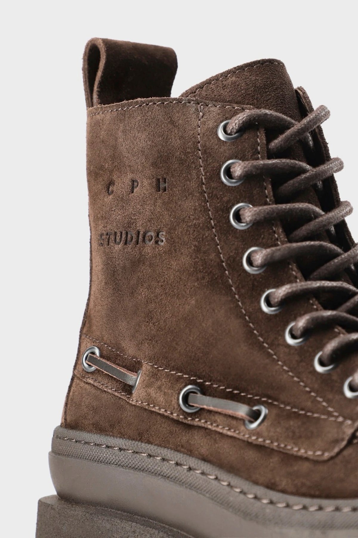 Close-up of a brown suede lace-up ankle boot with silver metal eyelets and a thick gray sole, shown against a light gray background.
