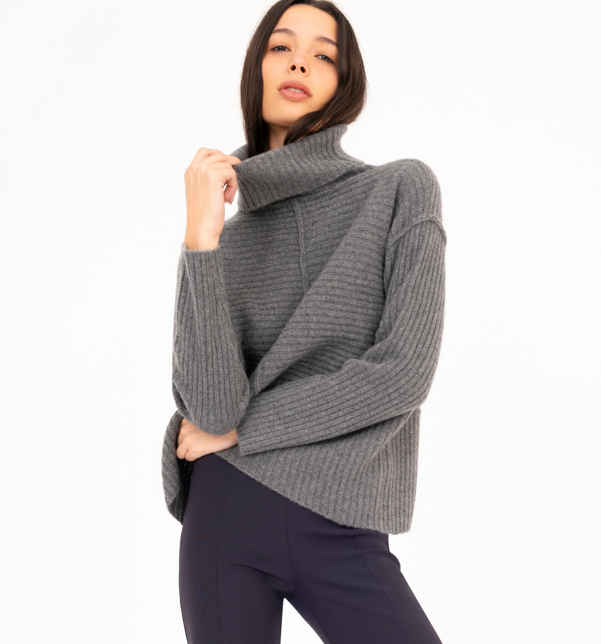 A woman wearing a ribbed gray turtleneck cashmere sweater and dark navy pants, posing against a clean white background.