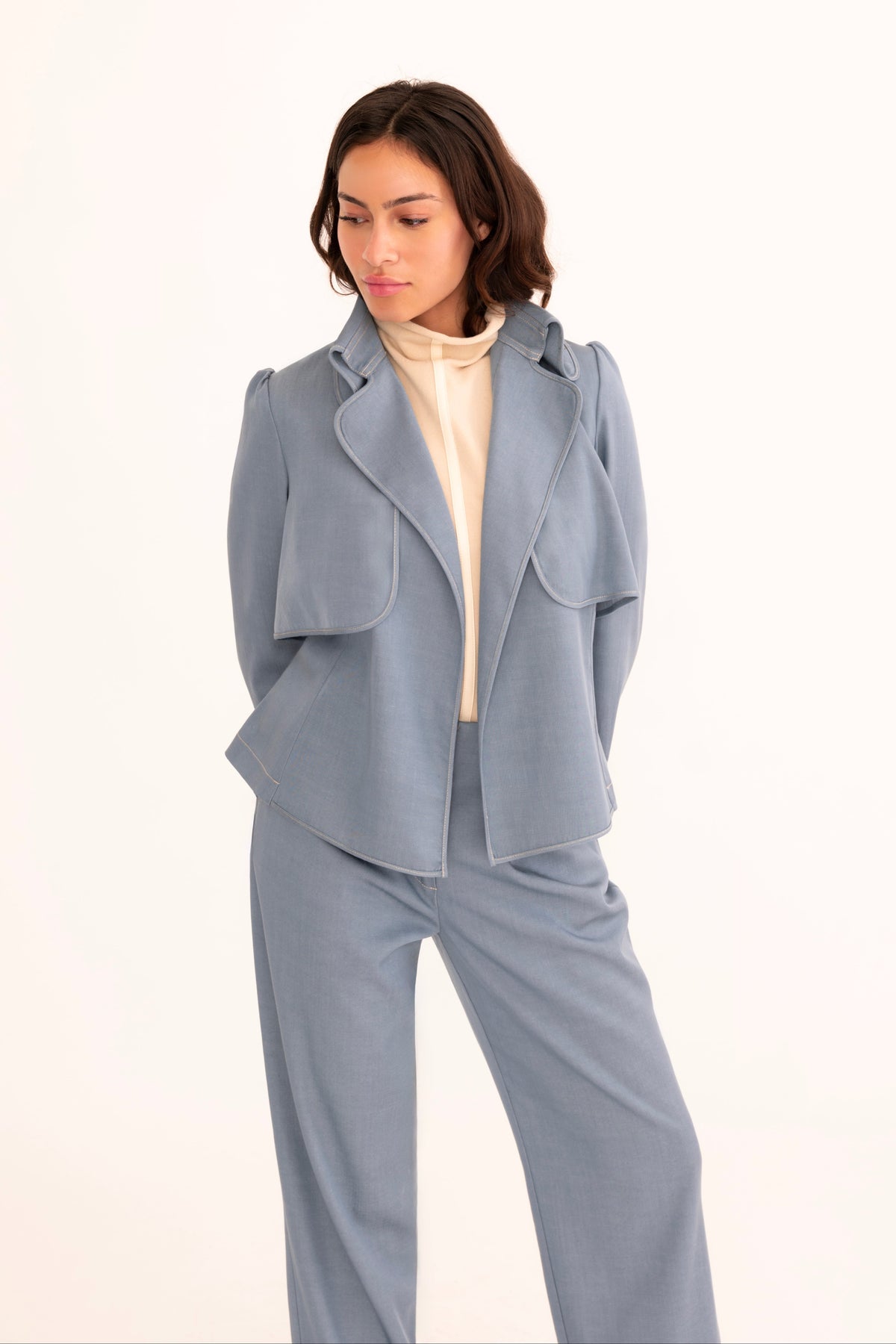 A model wearing a light blue tailored jacket and matching wide-leg trousers over a cream mock-neck top, standing against a plain white studio background.
