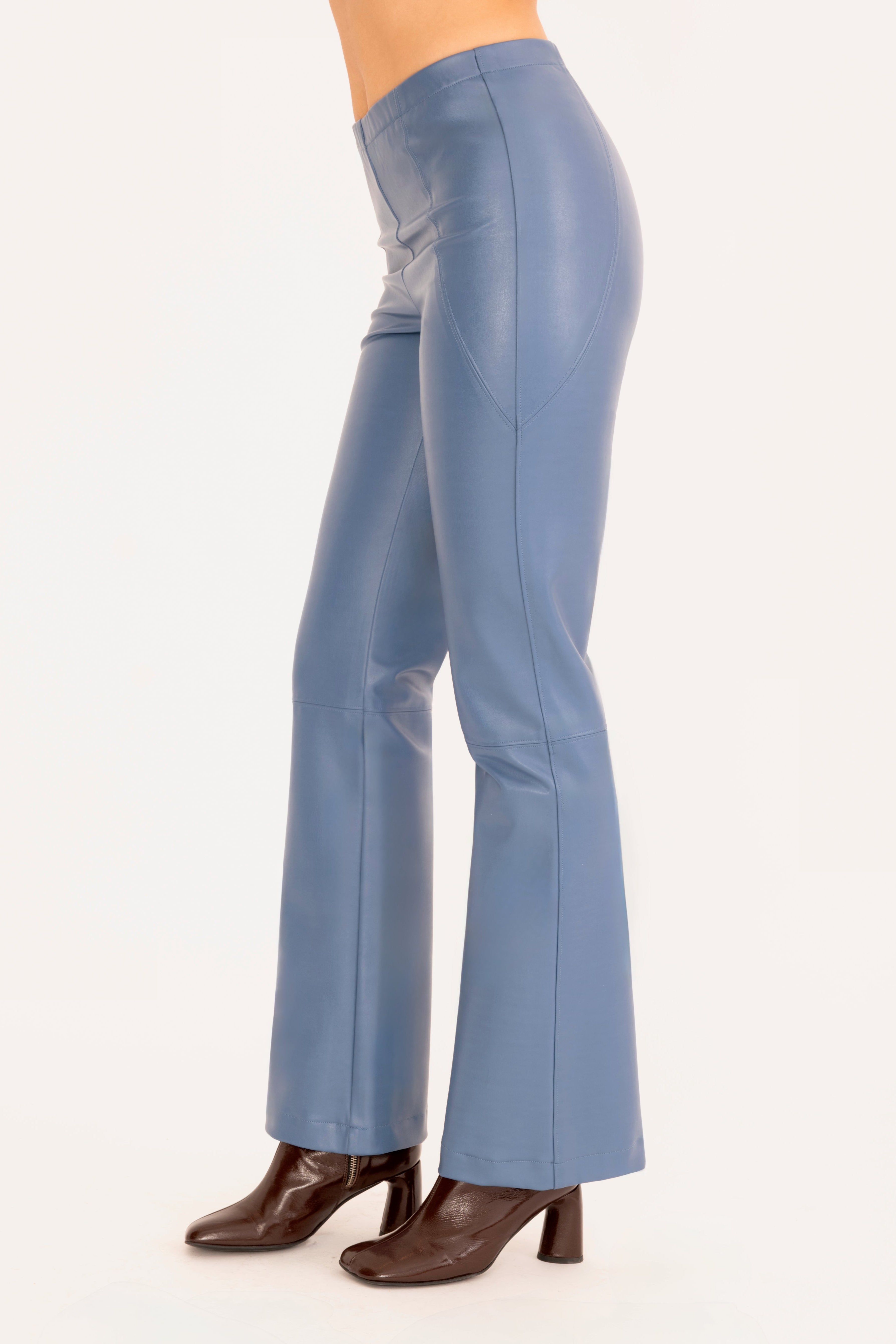 Side view of a model shown from the waist down wearing high-waisted, wide-leg blue faux-leather pants and dark brown heeled boots against a plain off-white background.