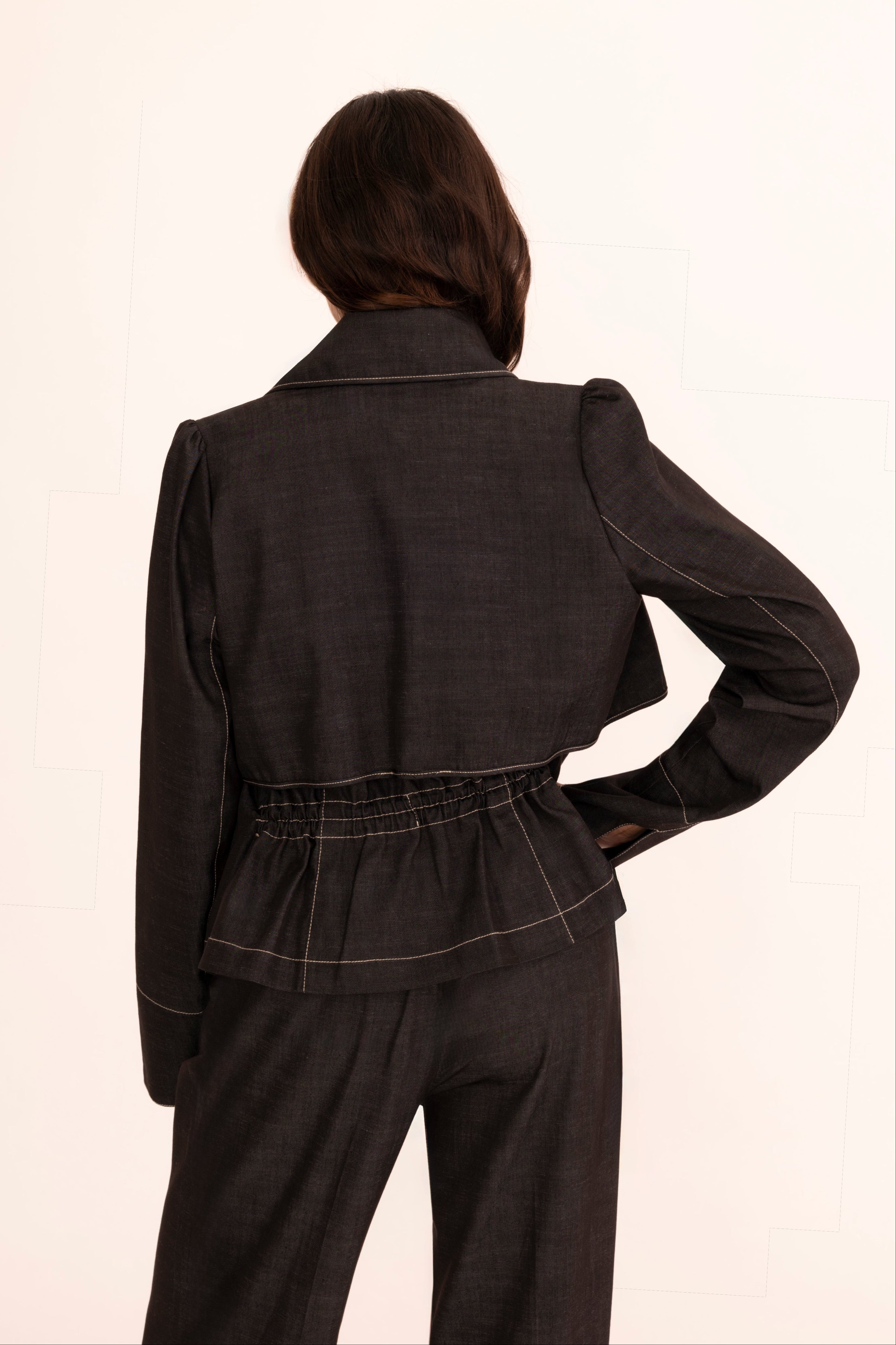 A woman modeling a light blue tailored denim suit with structured shoulders, flap pockets, and matching trousers, worn over a cream mock-neck top against a plain off-white studio background.