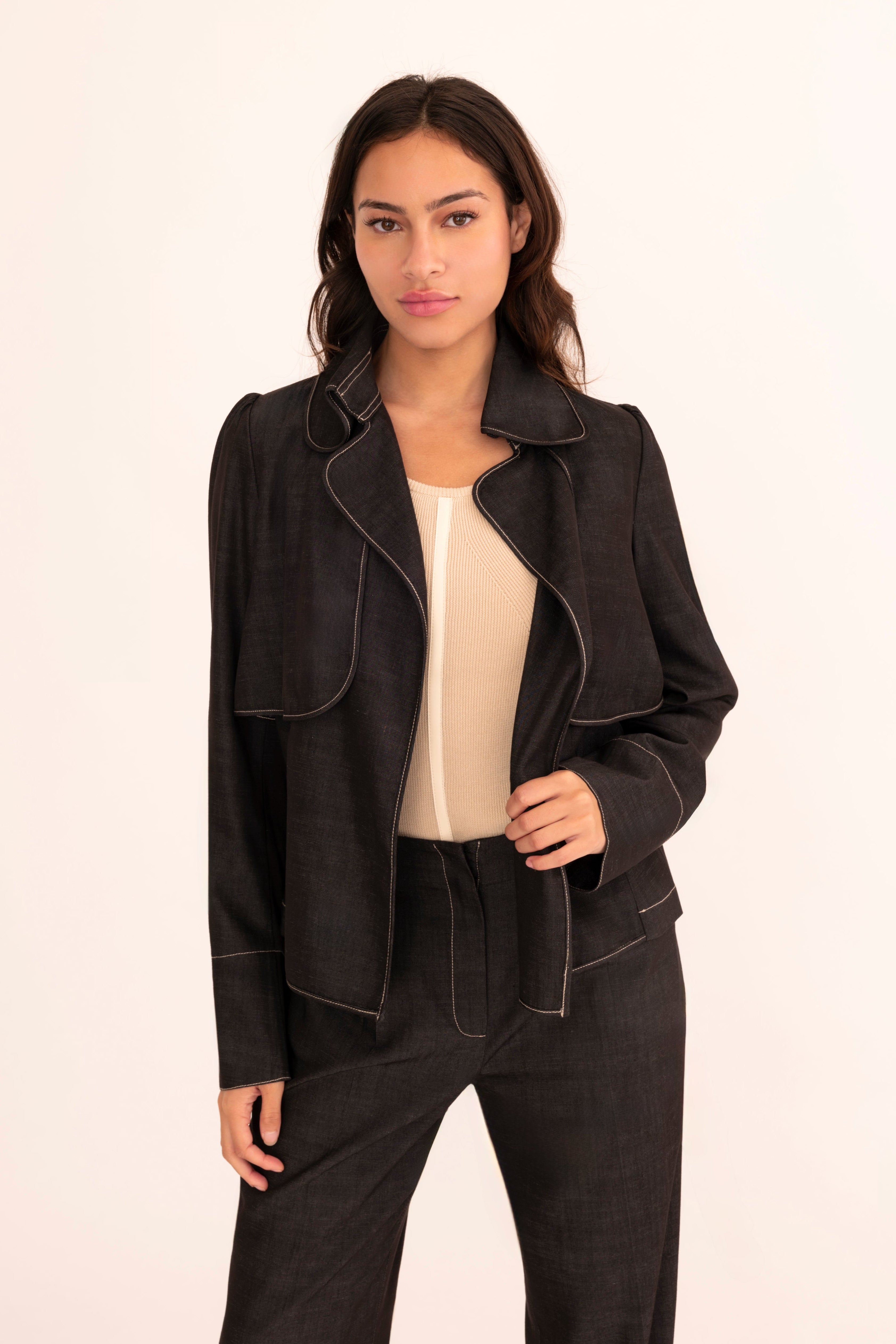 A woman modeling a black tailored denim suit with structured shoulders, flap pockets, and matching trousers, worn over a cream mock-neck top against a plain off-white studio background.