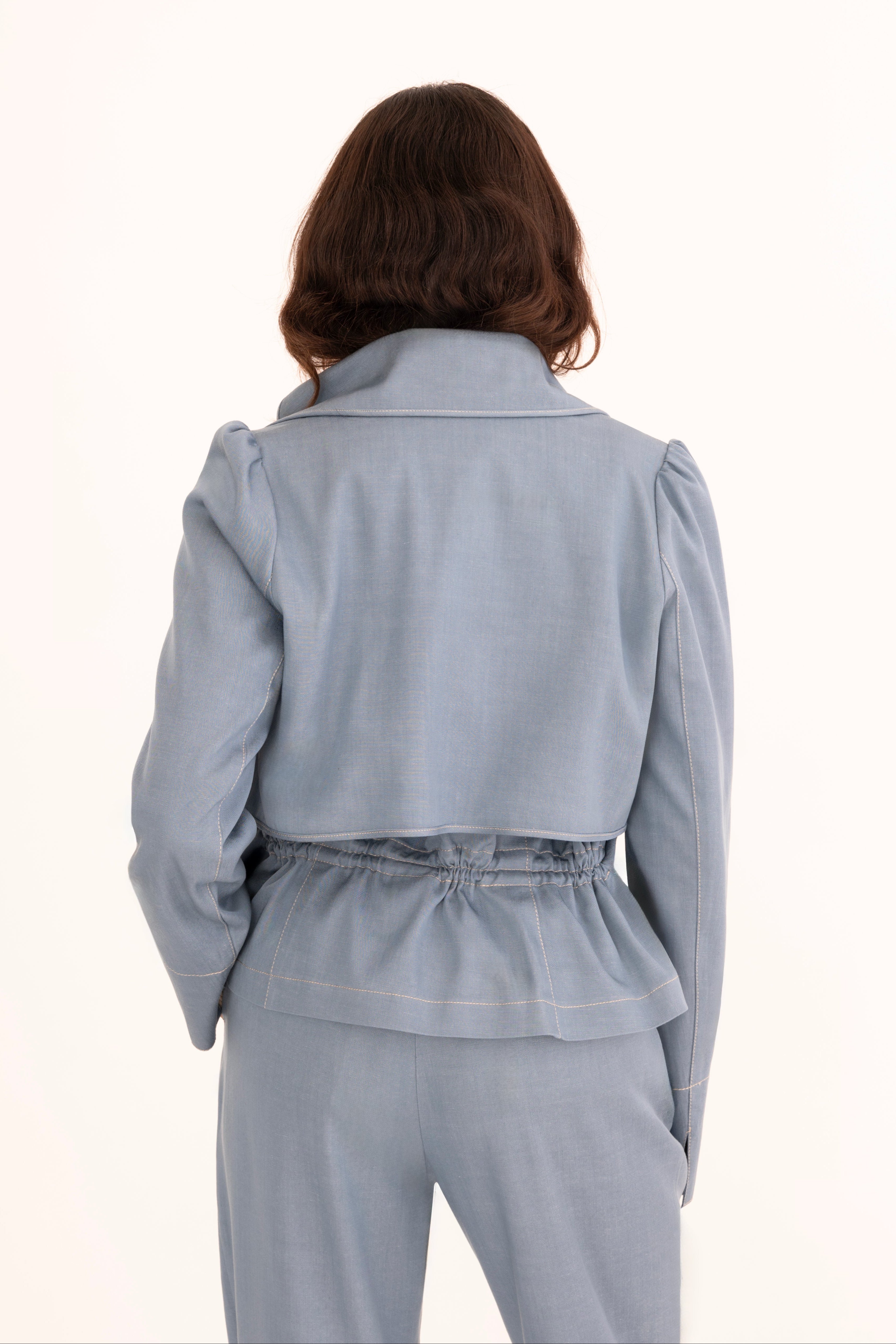 A woman modeling a light blue tailored denim suit with structured shoulders, flap pockets, and matching trousers, worn over a cream mock-neck top against a plain off-white studio background.