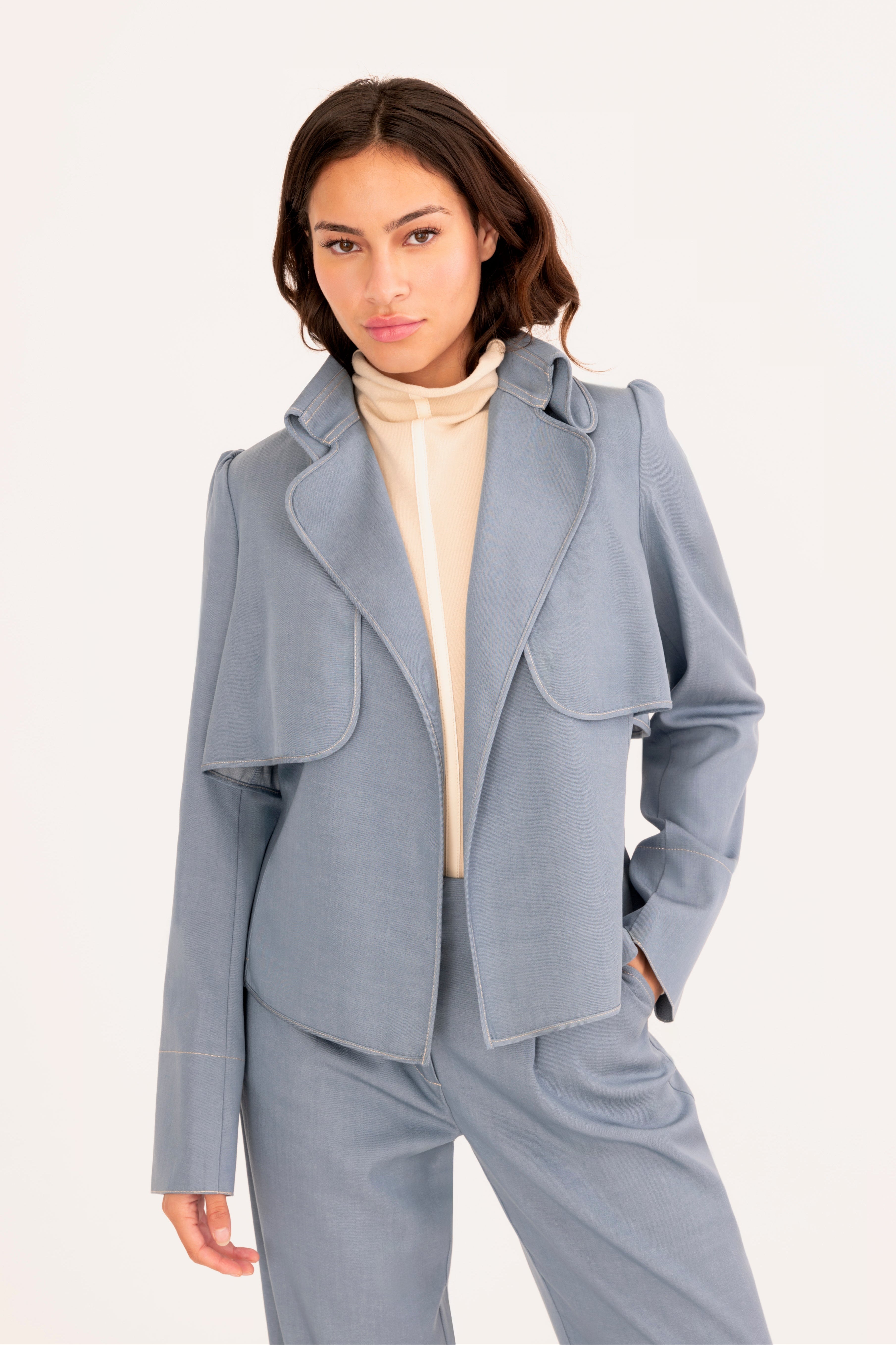 A woman modeling a light blue tailored denim suit with structured shoulders, flap pockets, and matching trousers, worn over a cream mock-neck top against a plain off-white studio background.