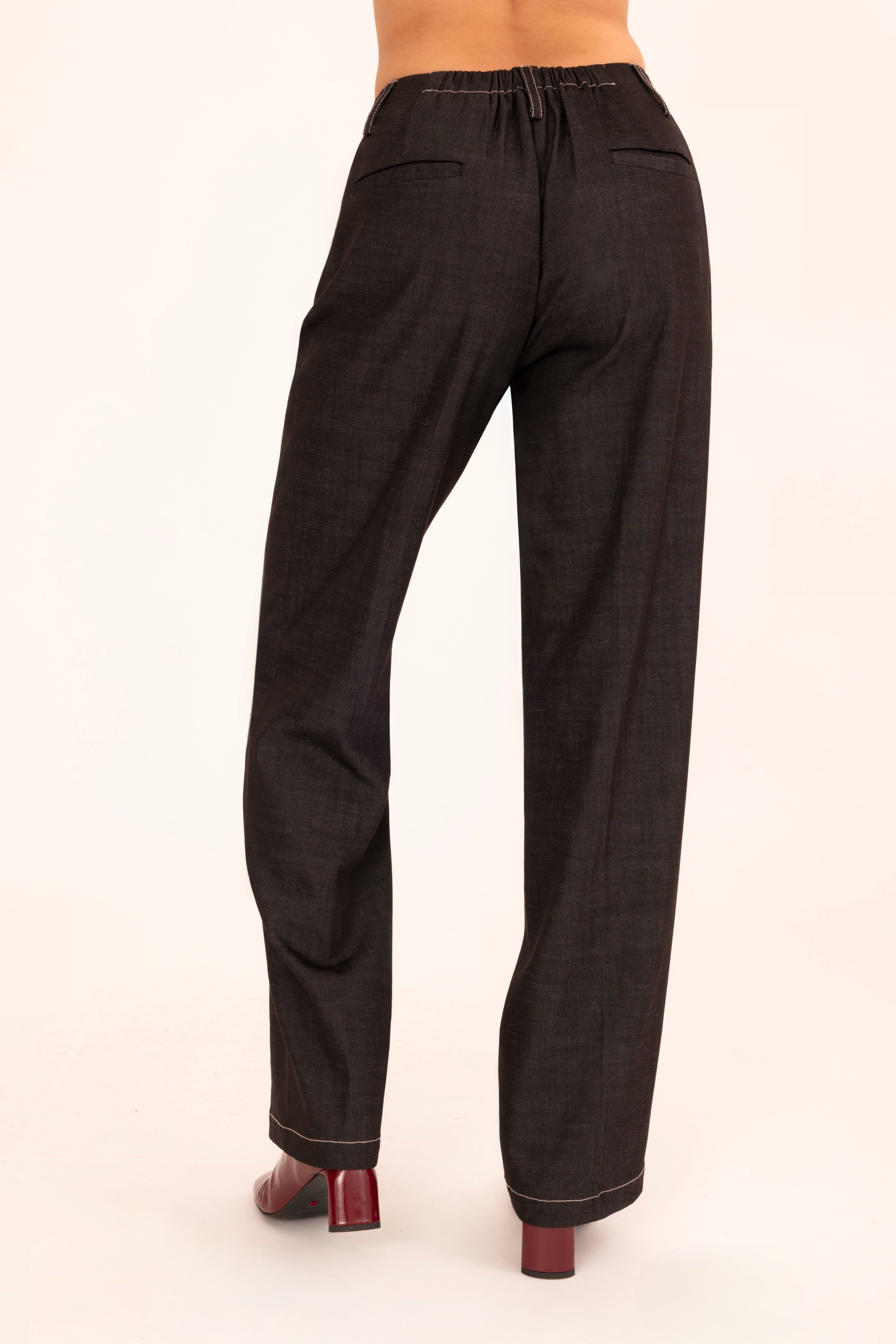 A model is shown from the waist down wearing black denim trousers and dark red shoes, standing against a plain off-white background.