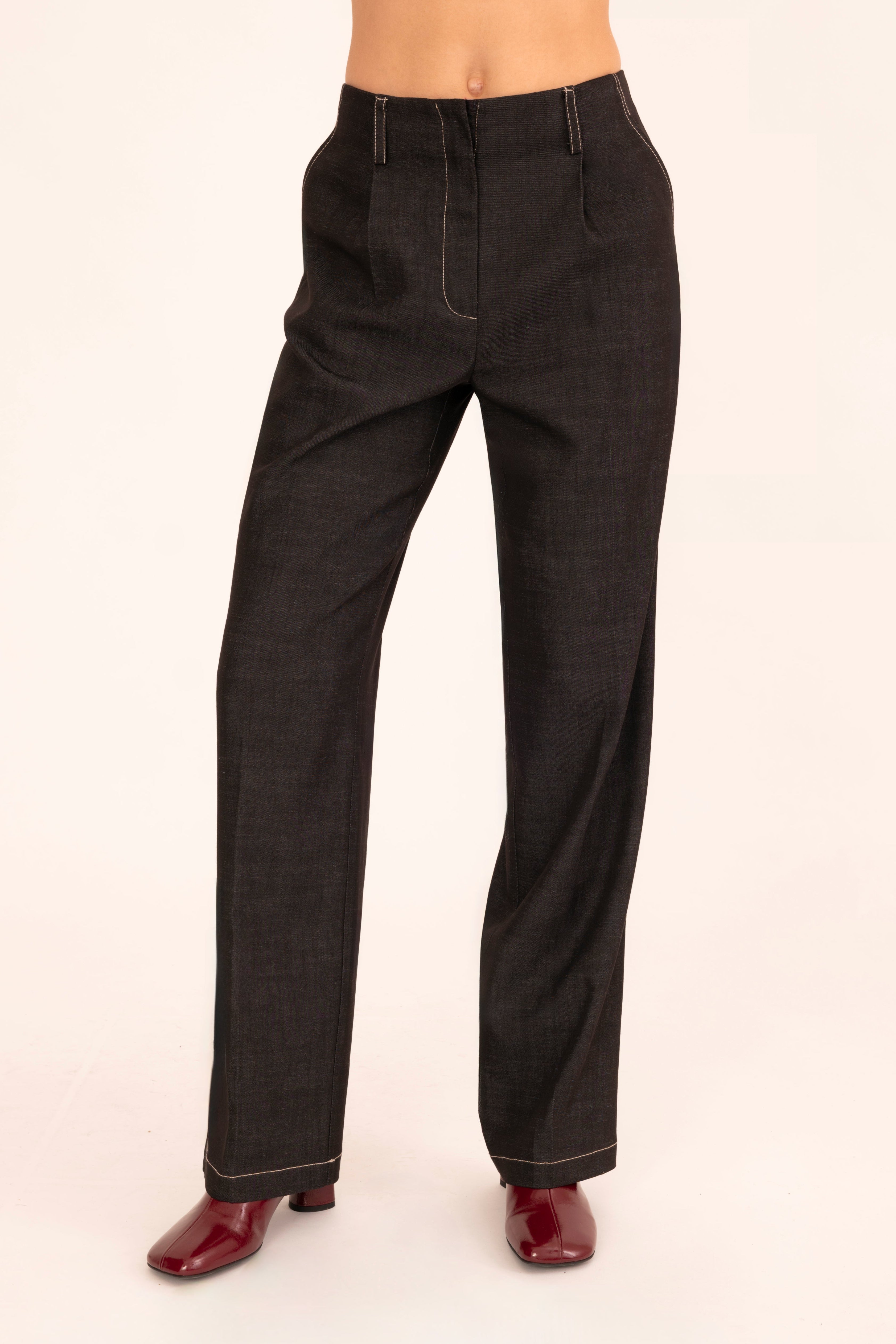 A model is shown from the waist down wearing black denim trousers and dark red shoes, standing against a plain off-white background.