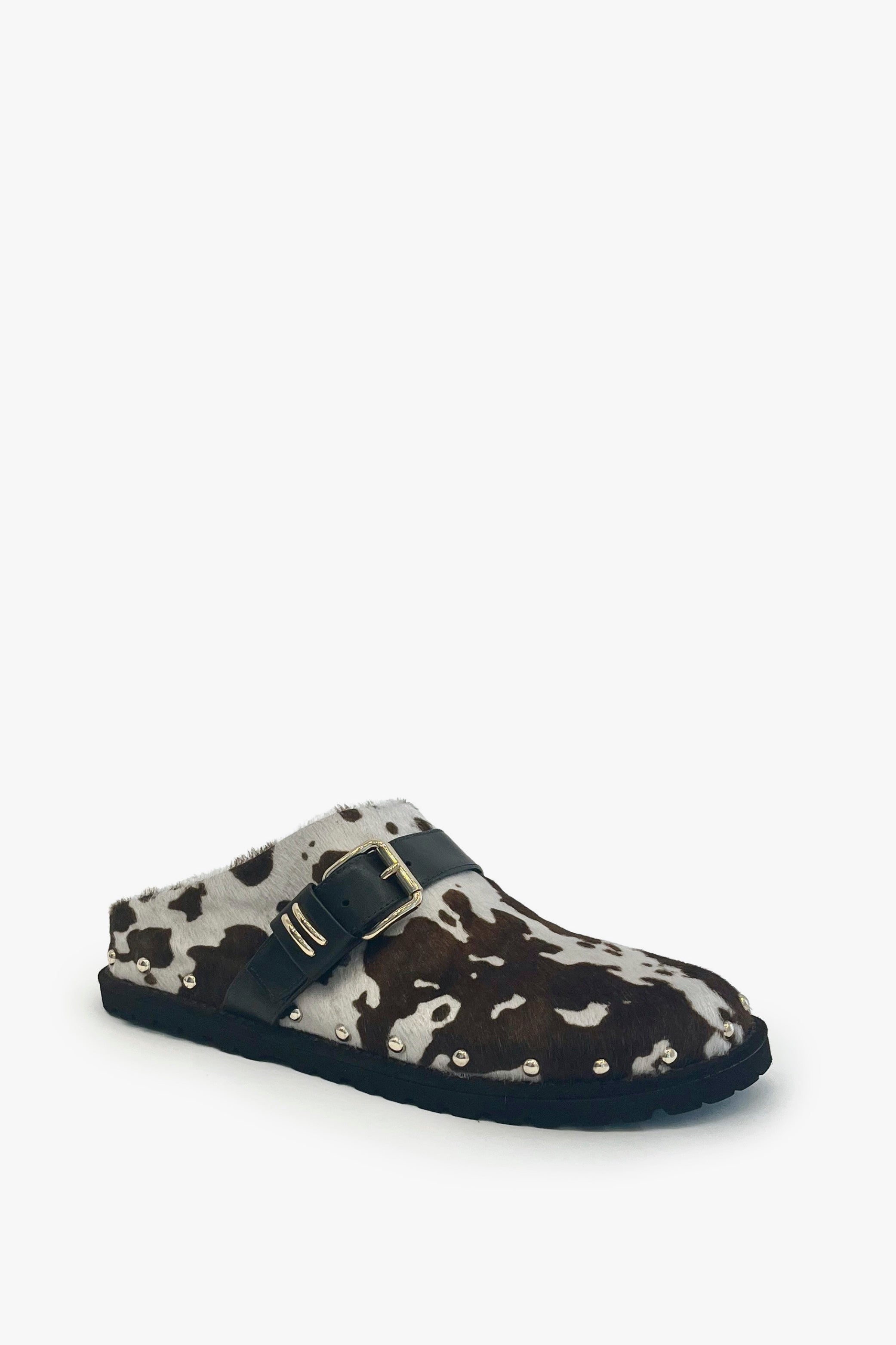 A single clog-style mule shoe with a brown and beige cowhide pattern, a black strap across the top, and a gold buckle, set against a plain white background.