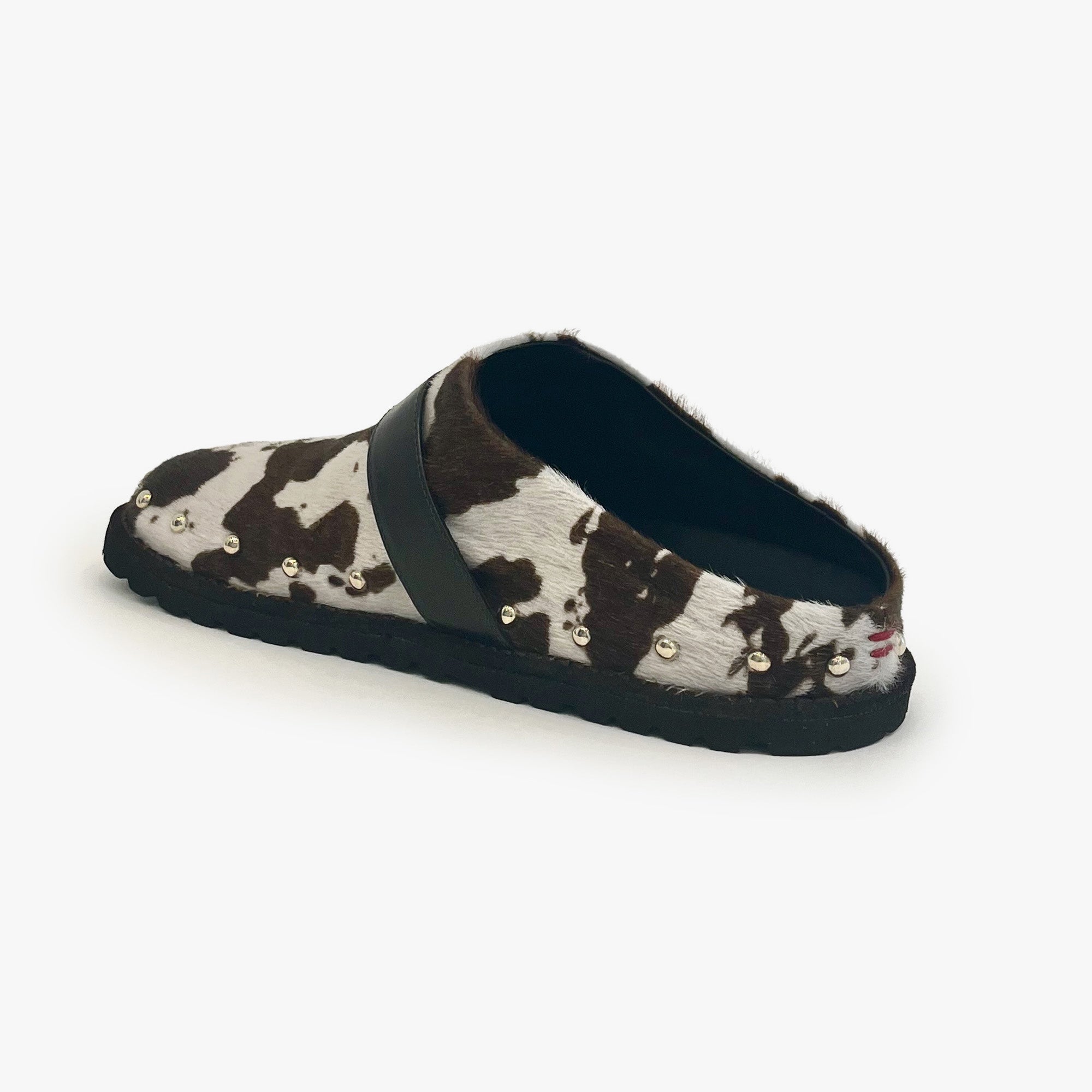 A single slip-on mule shoe featuring a brown and white cow print pattern, a wide black strap across the top, and small gold-tone studs along the side, set against a plain white background.