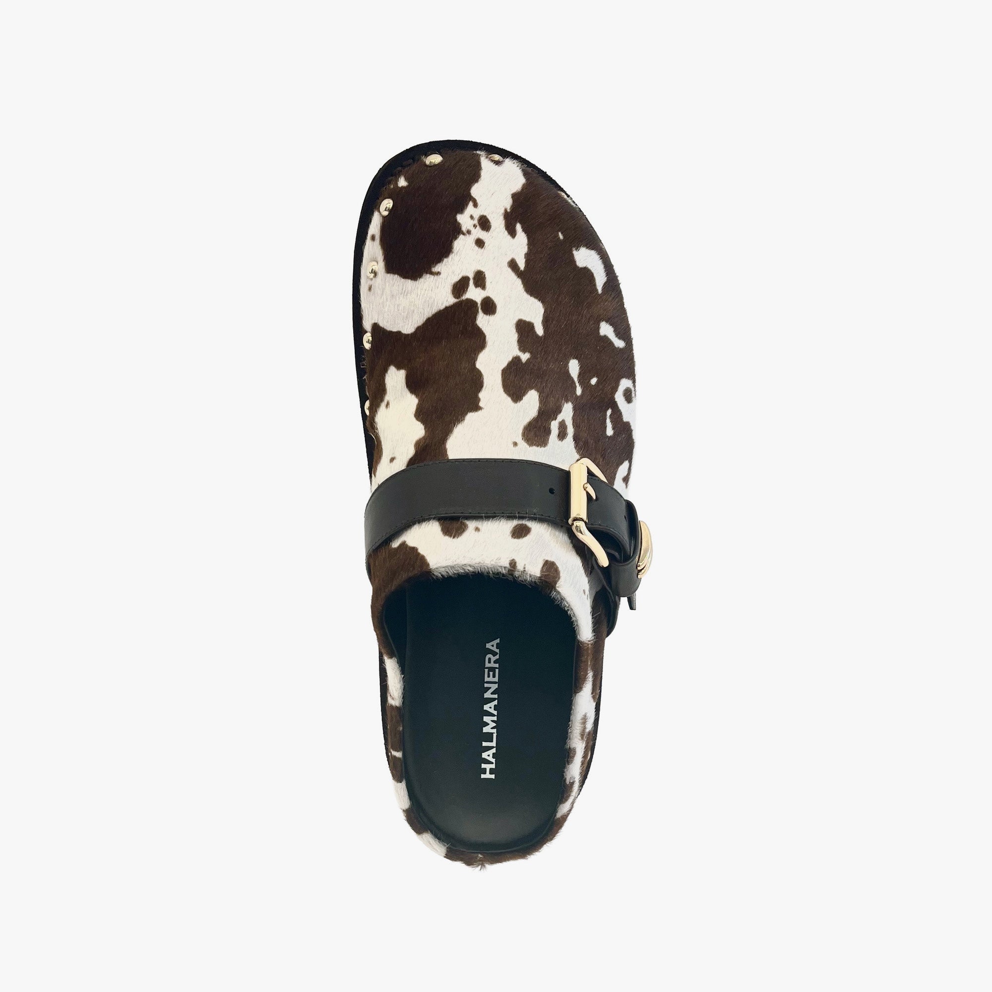 A single clog-style shoe shown from a top-down view on a light gray background. The upper is made of brown and white cowhide with a black strap and gold-tone buckle across the top.