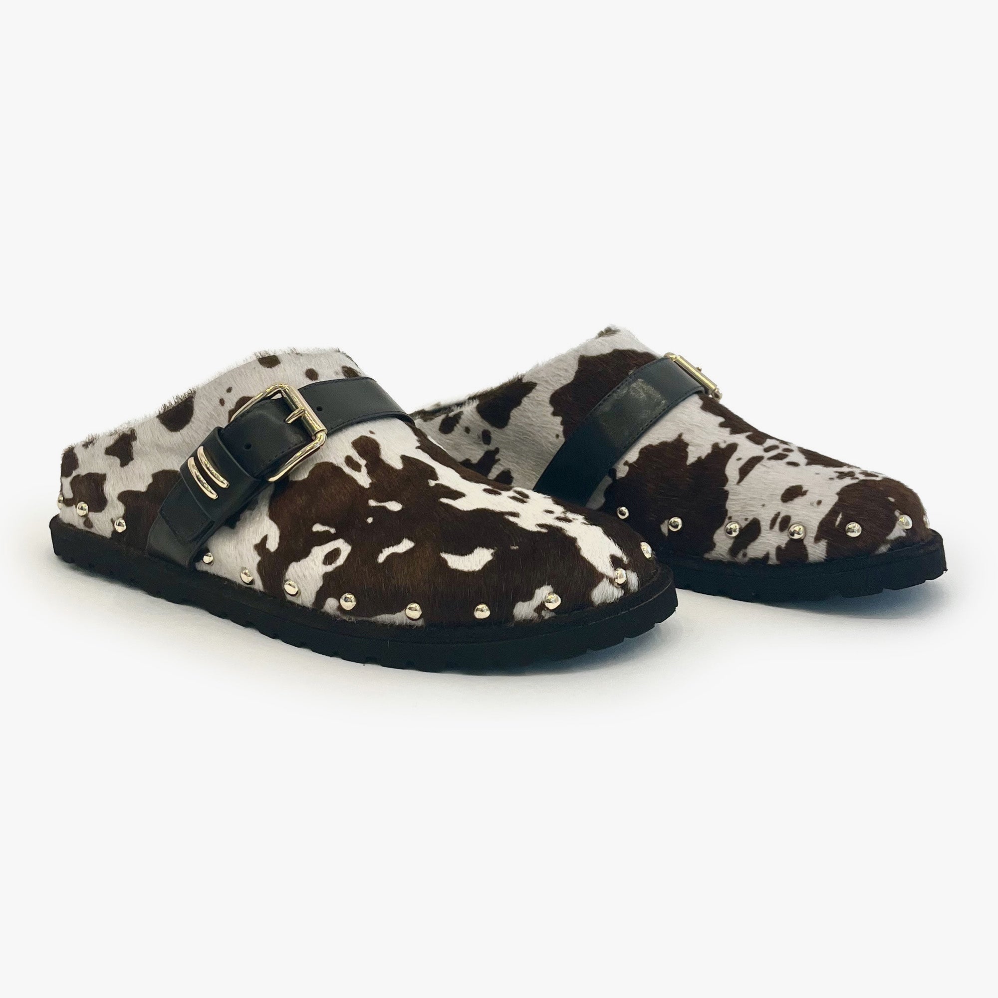 A pair of slip-on clogs with a brown and white cowhide pattern, featuring black straps with gold buckles and gold metal studs on the toe, set against a plain white background.