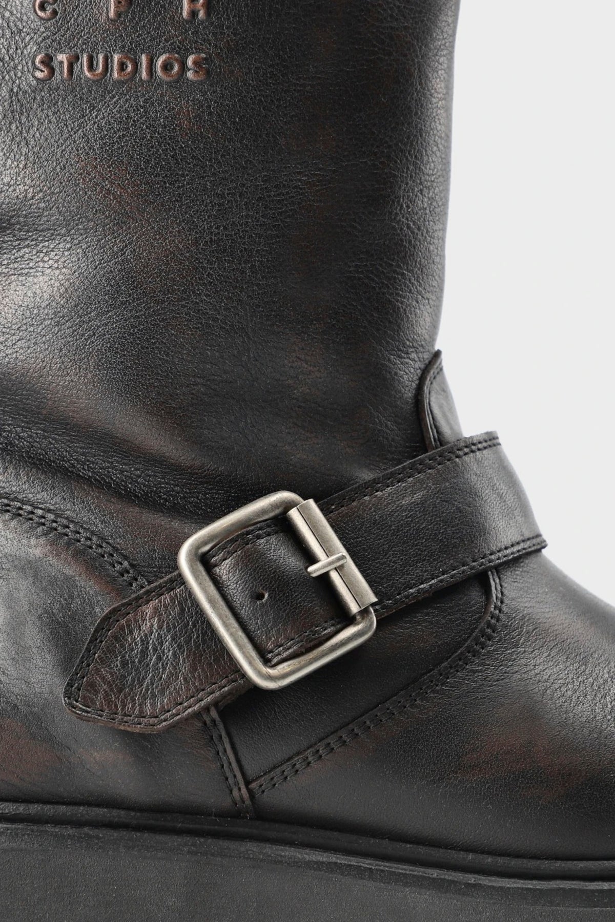 Close-up of a dark brown leather boot with a decorative strap and silver buckle, showing part of the embossed 'CPR STUDIOS' branding on the shaft against a light gray background.