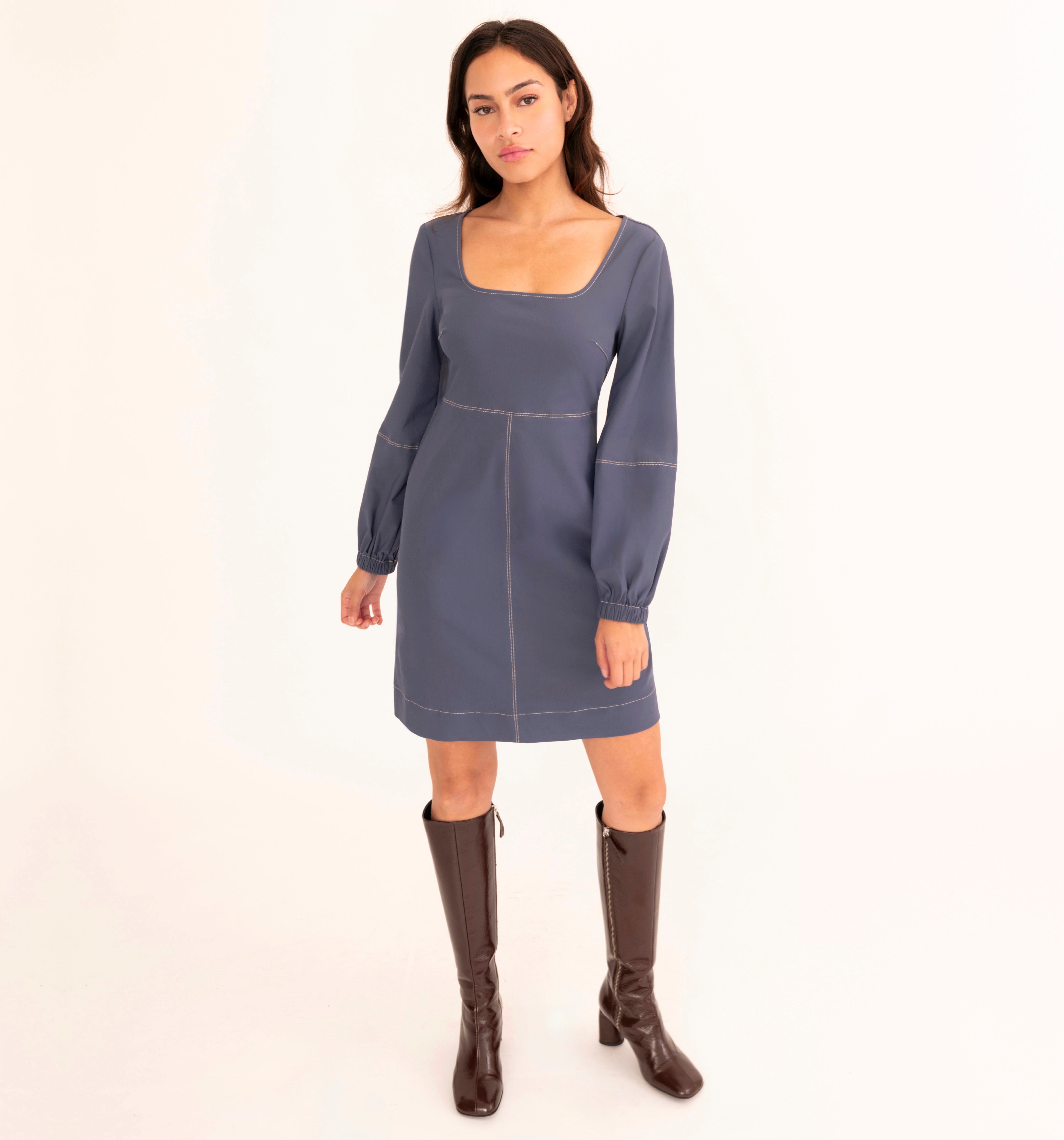 A woman modeling a slate blue long-sleeve mini dress with a square neckline and dark brown knee-high leather boots, standing against a plain white studio background.