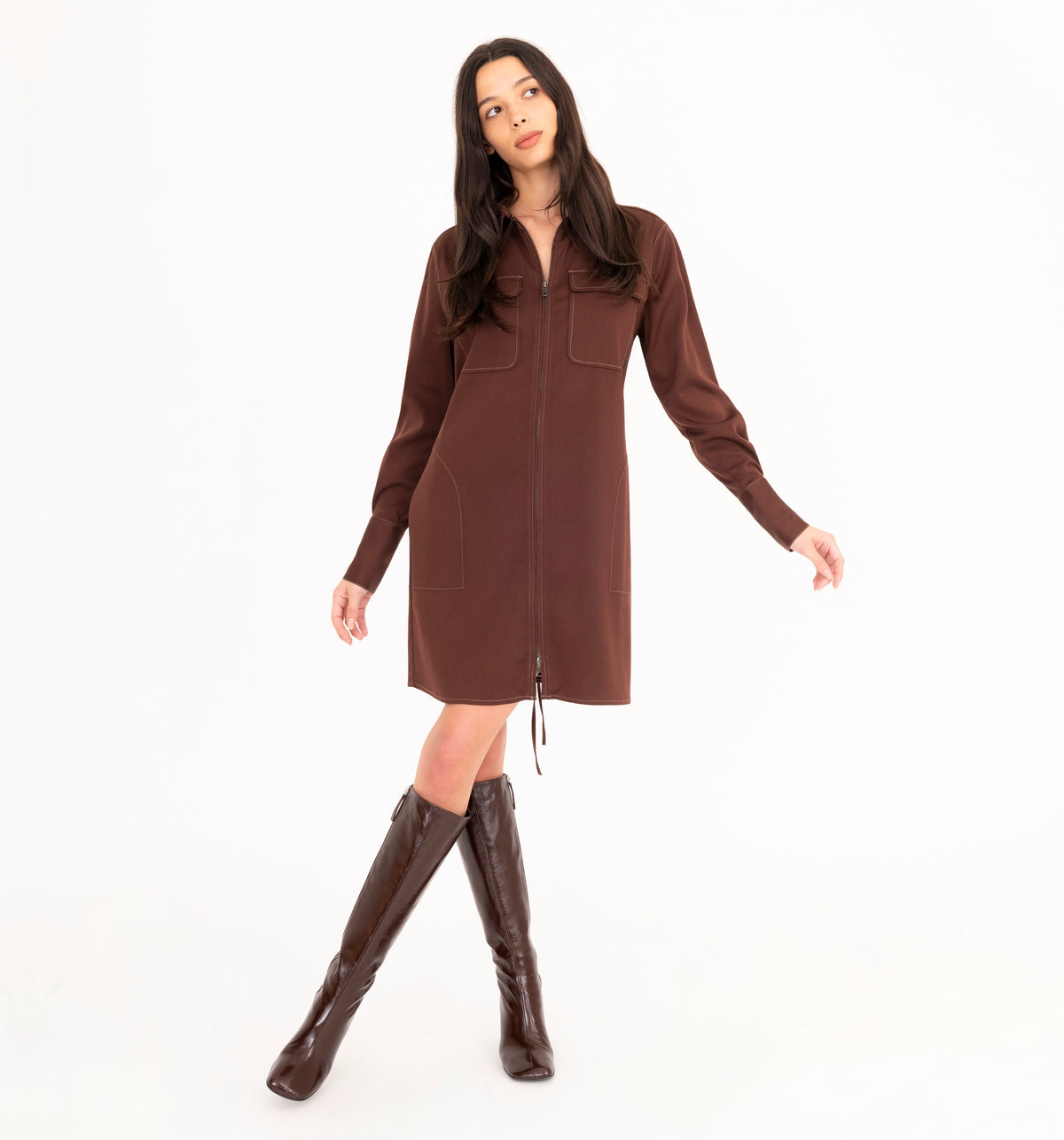 A woman modeling a brown zip-front denim dress with long sleeves and front pockets, paired with matching brown knee-high boots, against a plain white studio background.