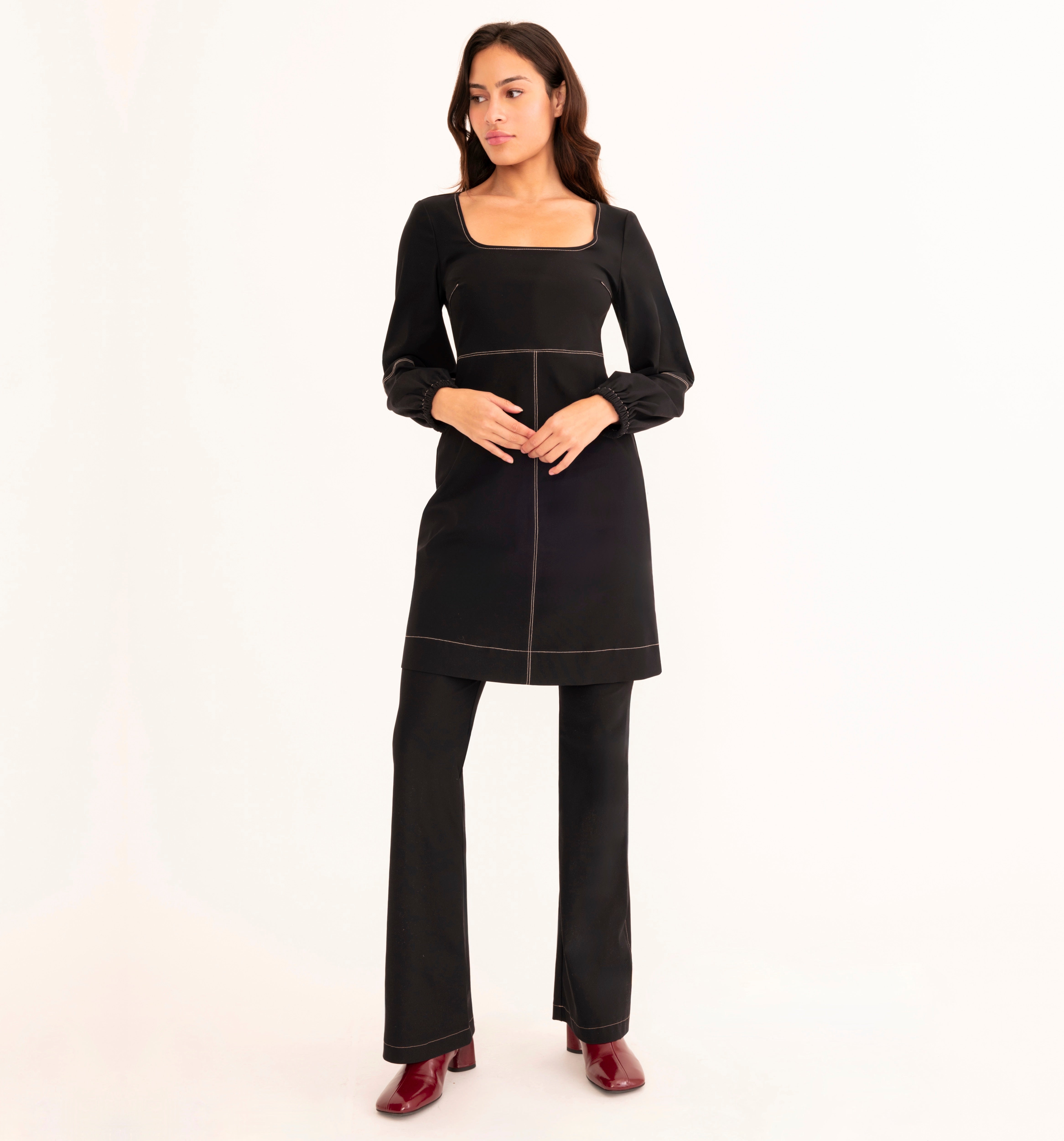 A woman modeling a black long-sleeve tunic dress with contrasting light piping, paired with black wide-leg pants and burgundy shoes, against a plain white studio background.