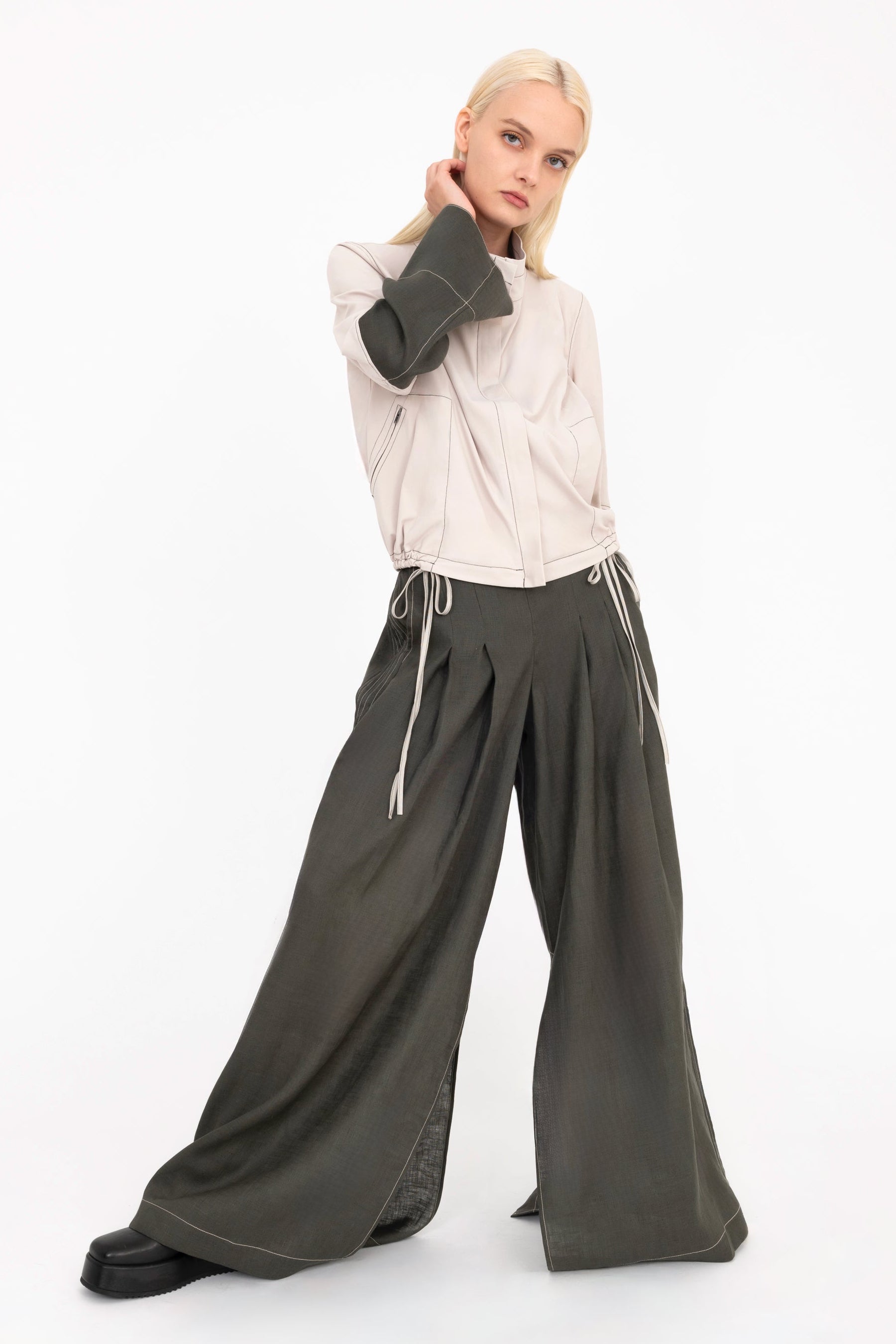 Woman wearing a beige jacket and  pine green pants with a white background