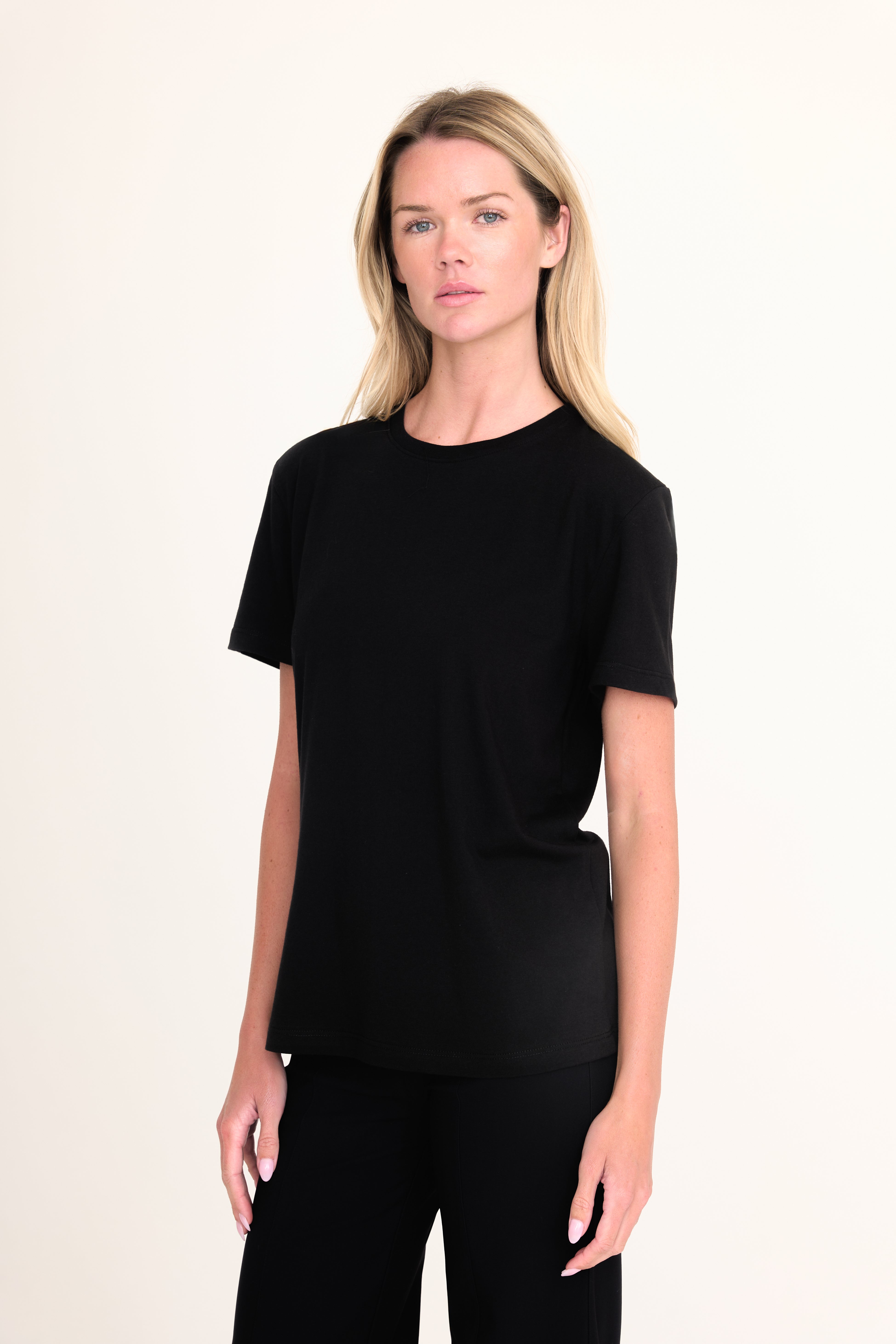 A blonde woman modeling a black short-sleeve crewneck t-shirt and black pants against a plain off-white studio background.