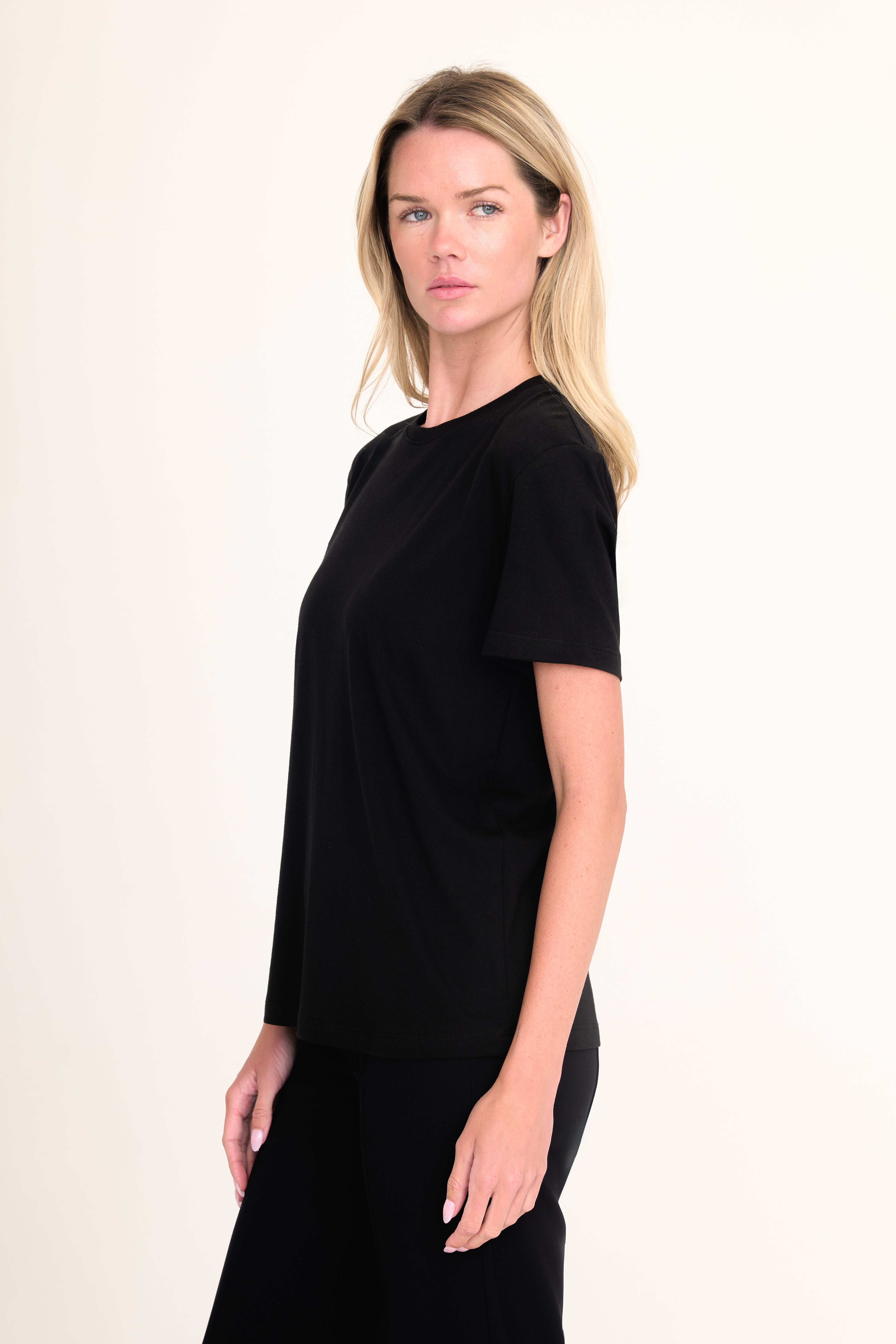A blonde woman modeling a black short-sleeve crewneck top against a plain off-white studio background.