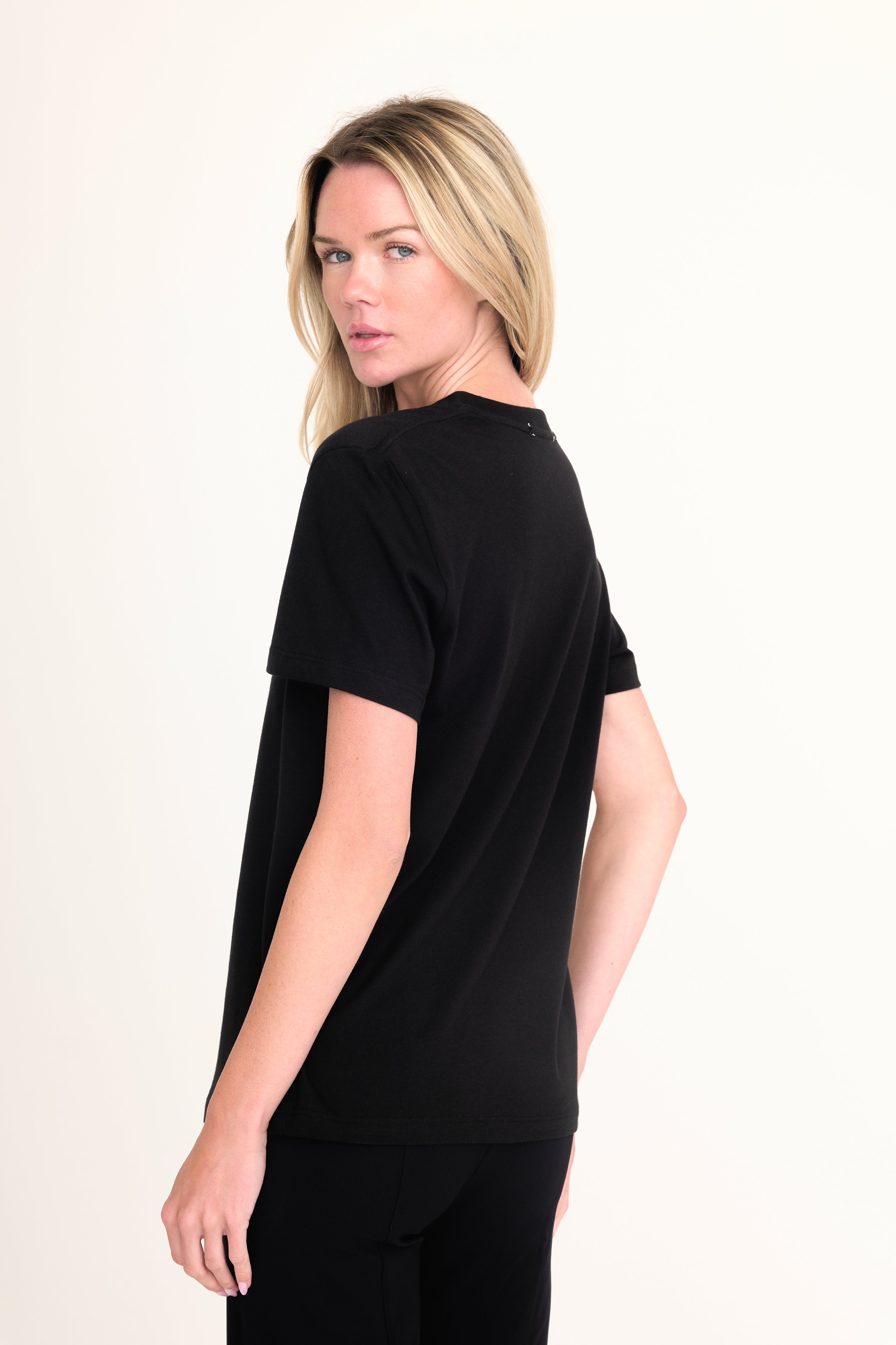 A blonde woman wearing a black short-sleeve crewneck t-shirt, shown from a three-quarters back view against a plain off-white studio background.