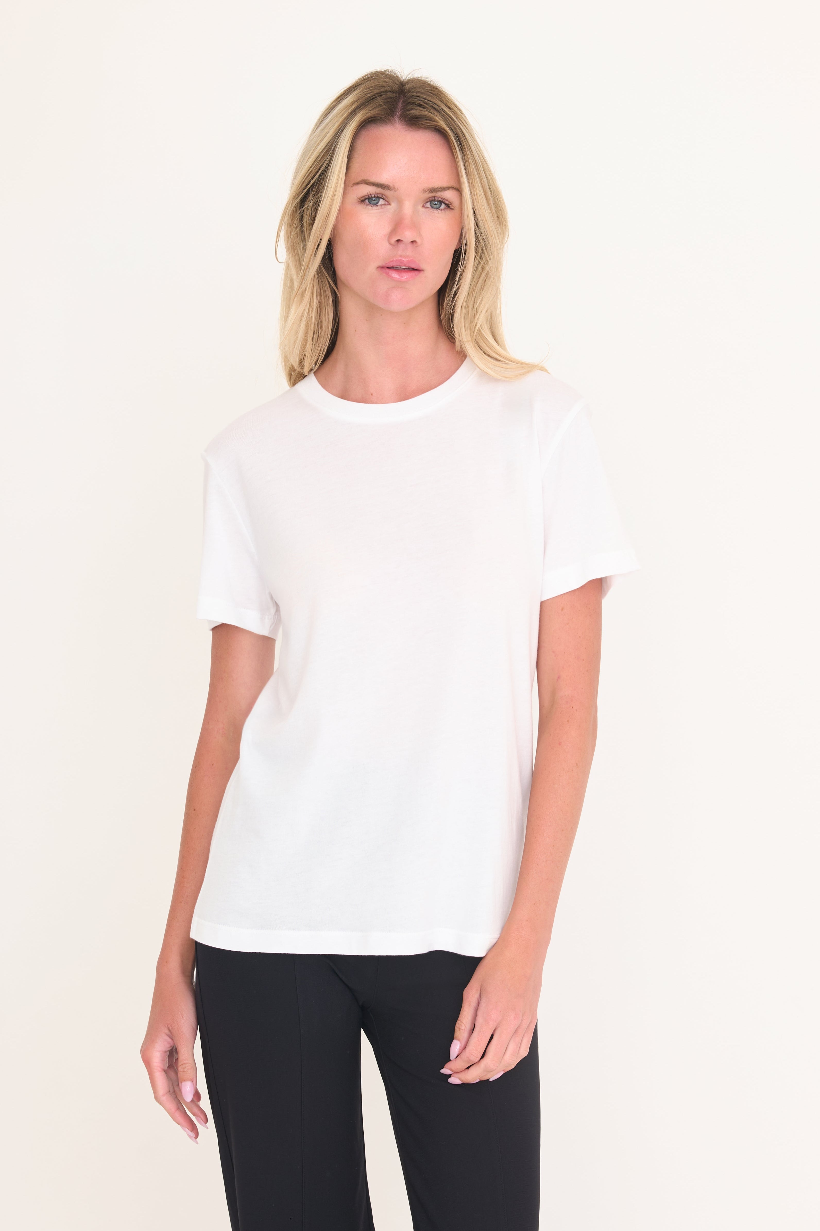A blonde woman modeling a plain white short-sleeve crewneck t-shirt paired with black pants, standing against a solid off-white studio background.