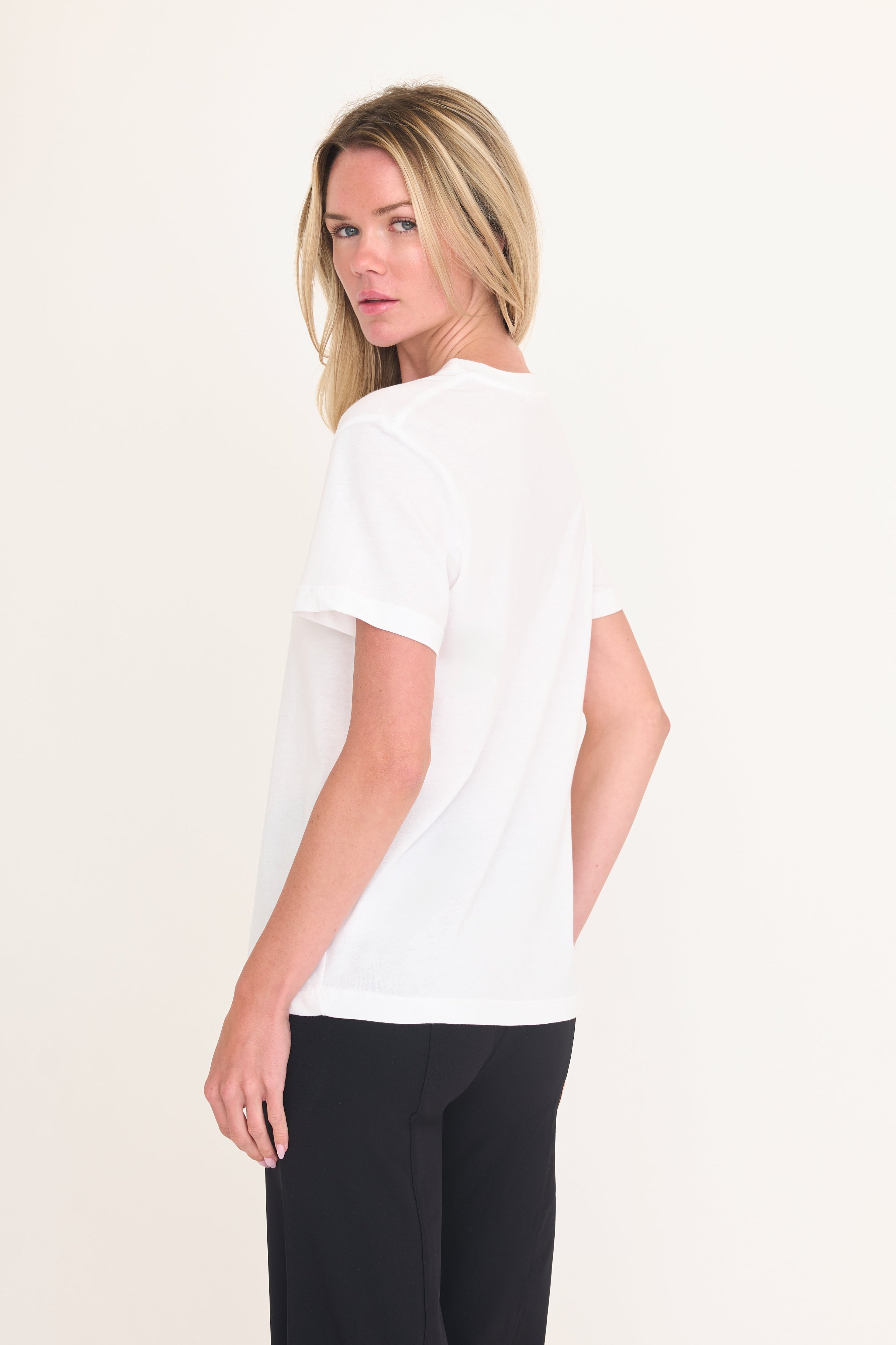 A blonde woman shown from the back wearing a white short-sleeve crewneck t-shirt and black pants, standing against a plain off-white studio background.