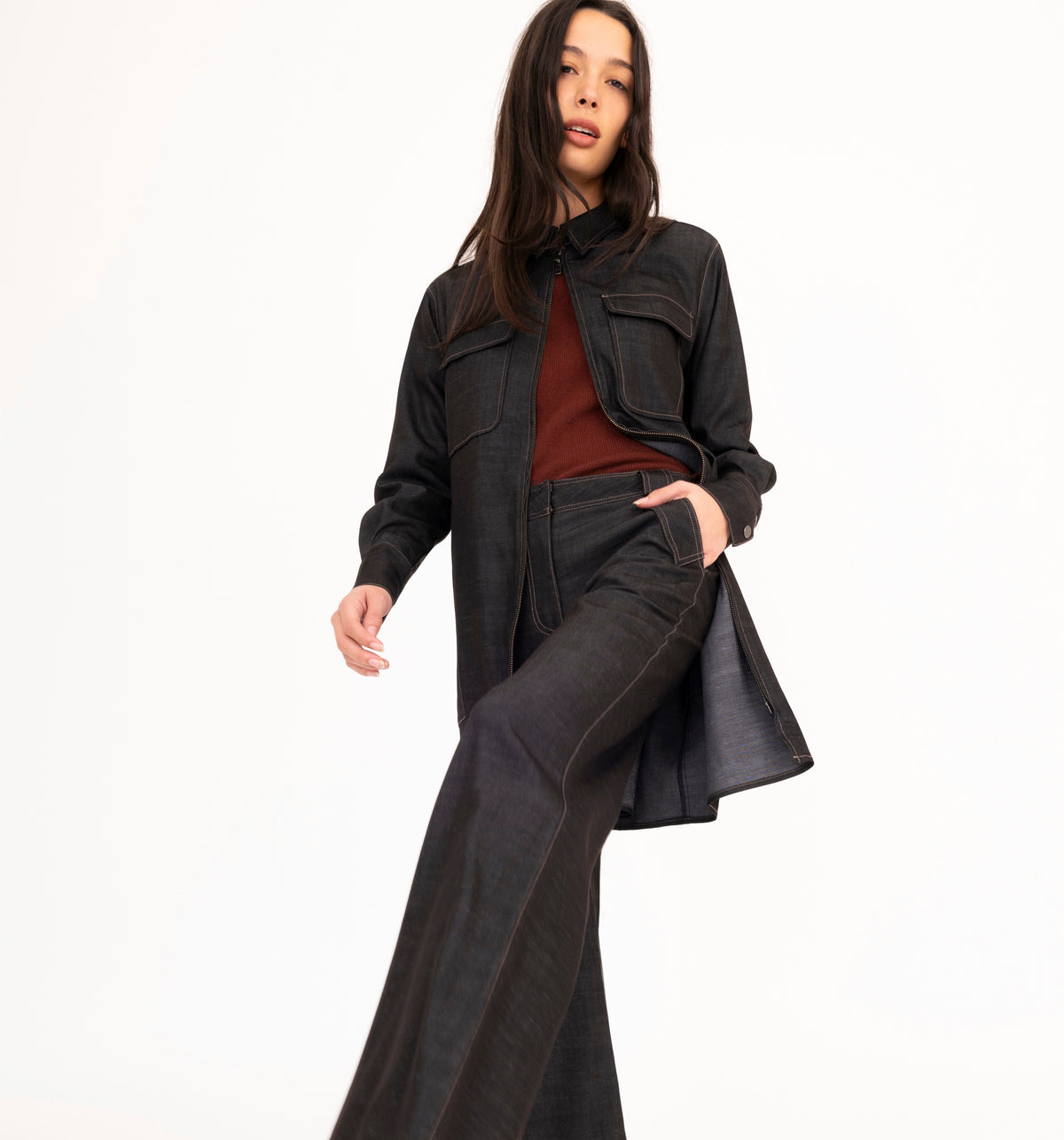 A model wearing a black denim jacket, matching wide-leg jeans, and a rust-colored sweater, posing against a solid white studio background.