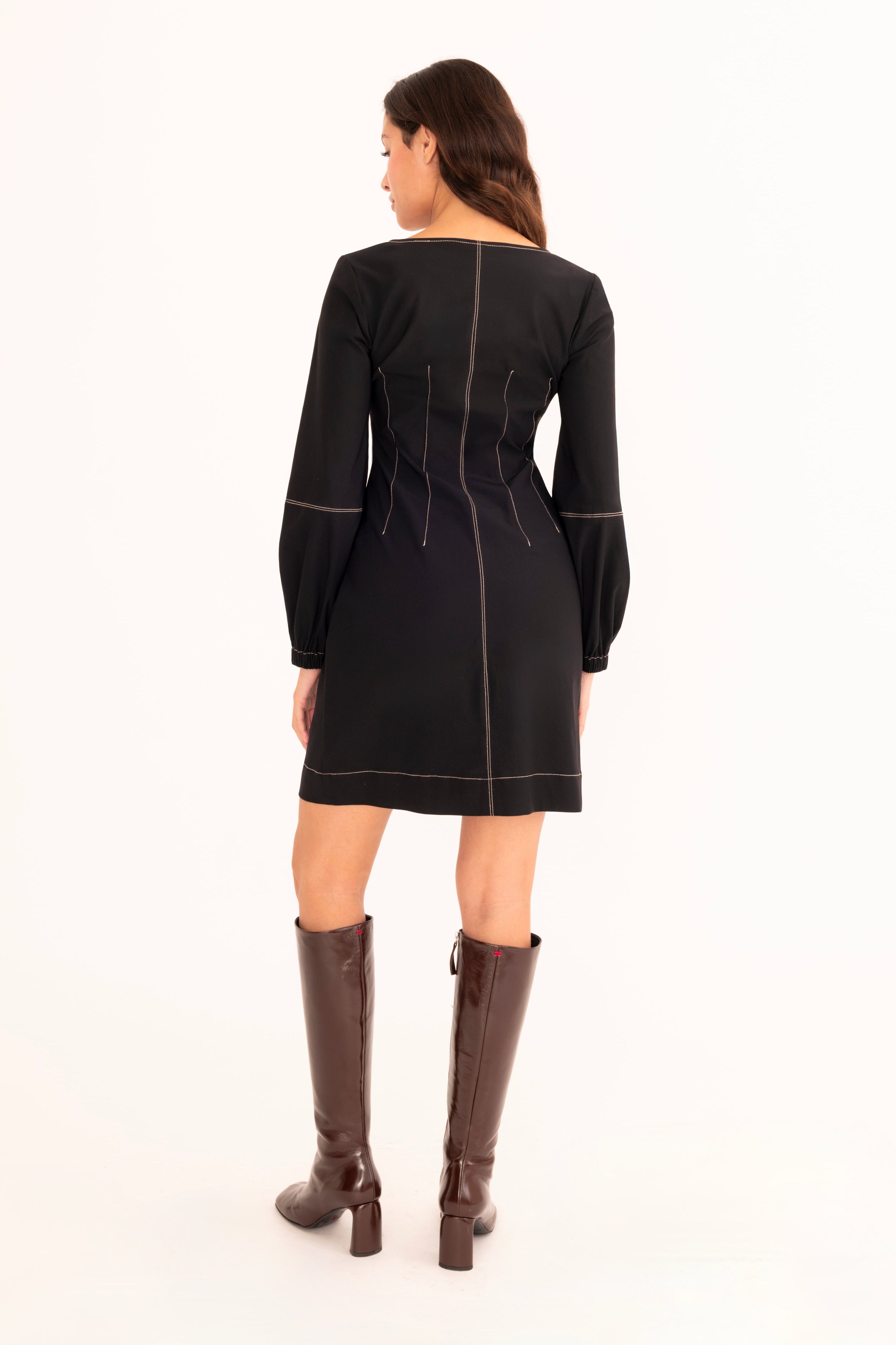 A woman viewed from the back wearing a black long-sleeve mini dress with contrast stitching and brown knee-high boots, standing against a plain white studio background.