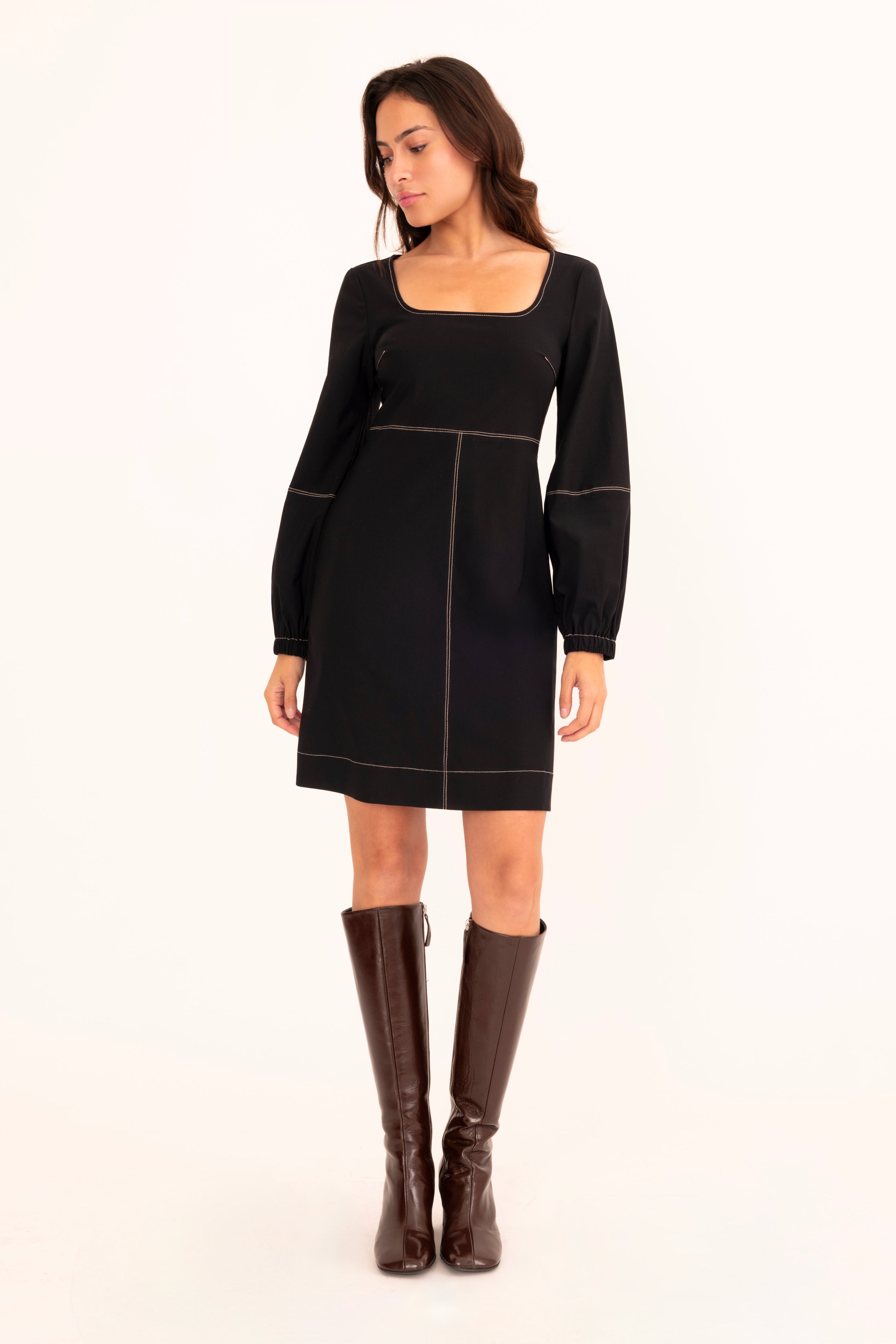 A woman modeling a black long-sleeve mini dress with contrast light piping and a square neckline, paired with dark brown knee-high boots, against a plain white studio background.