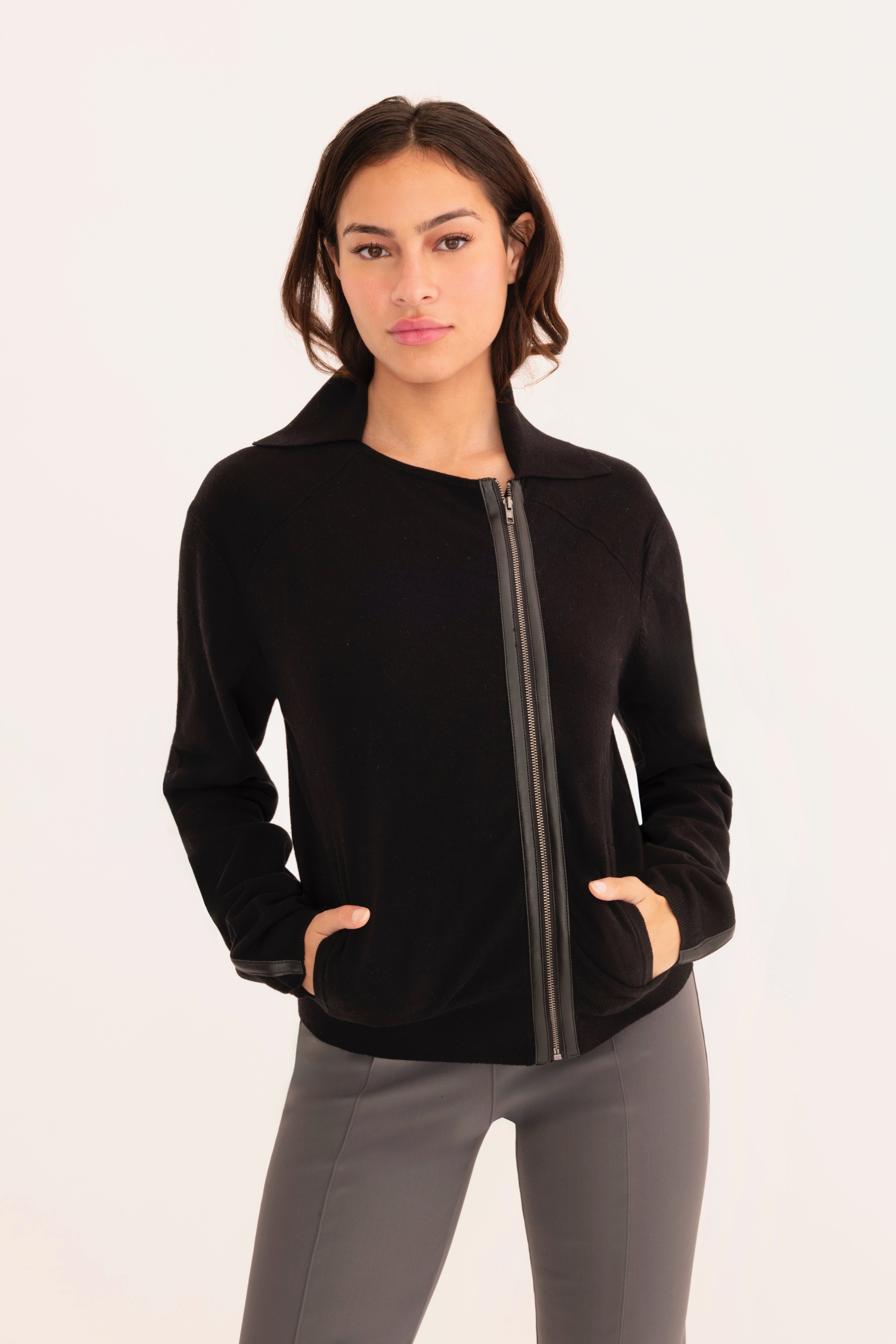 Front view of woman wearing a   black zip-up jacket and gray pants on a white background