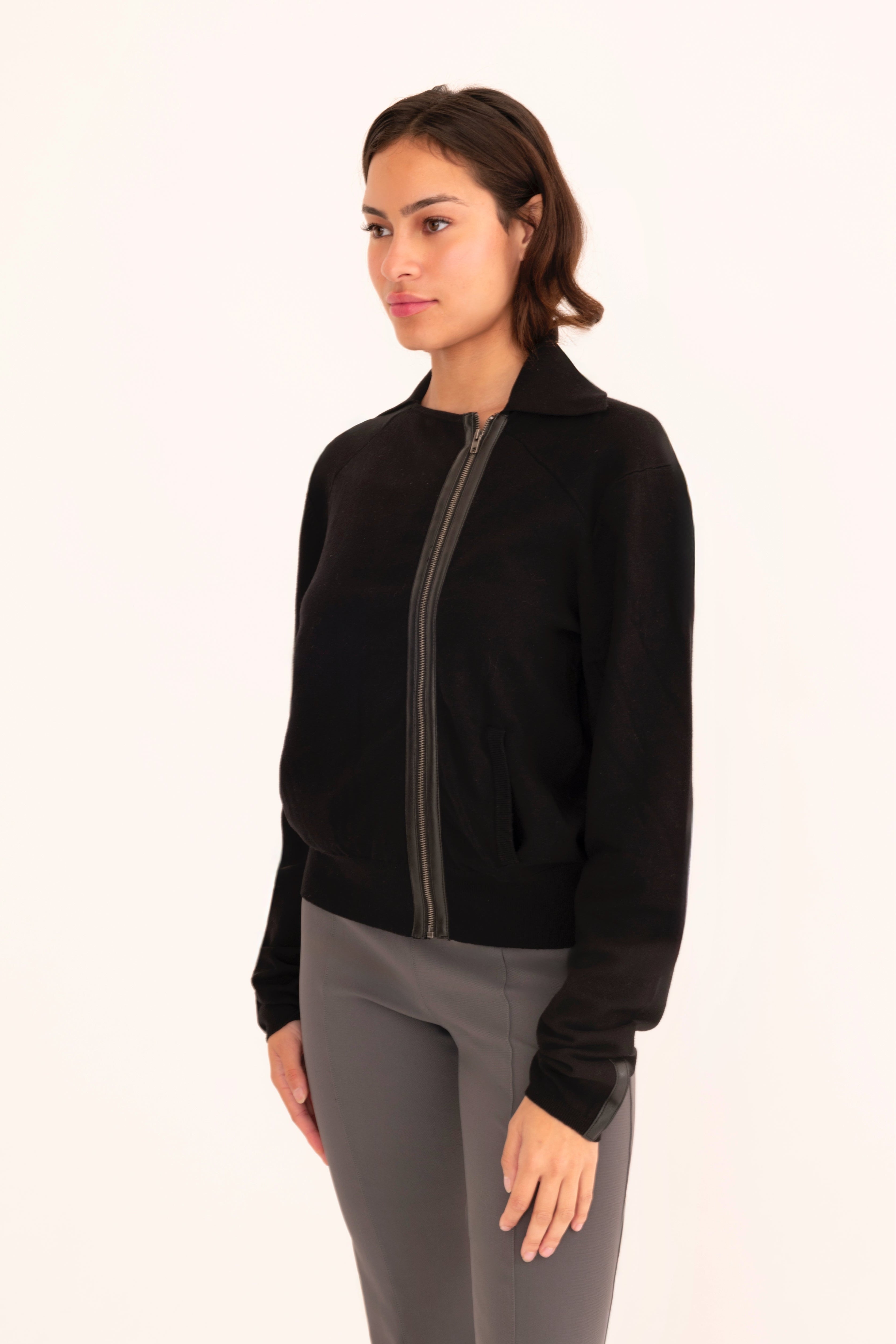 Side view of woman wearing a   black zip-up jacket and gray pants on a white background