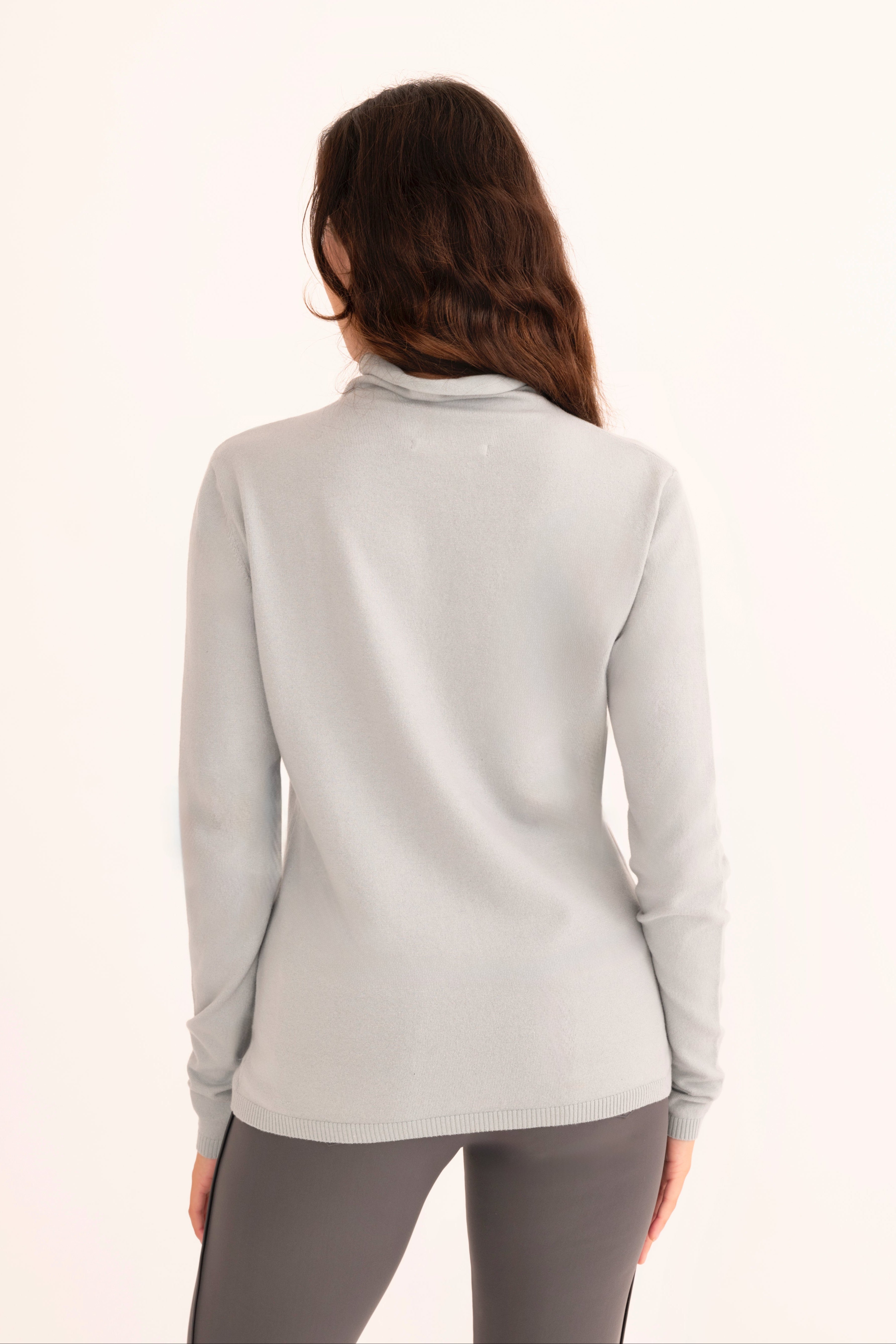 Back view of a woman wearing a light blue funnel neck top and gray pants on a white background