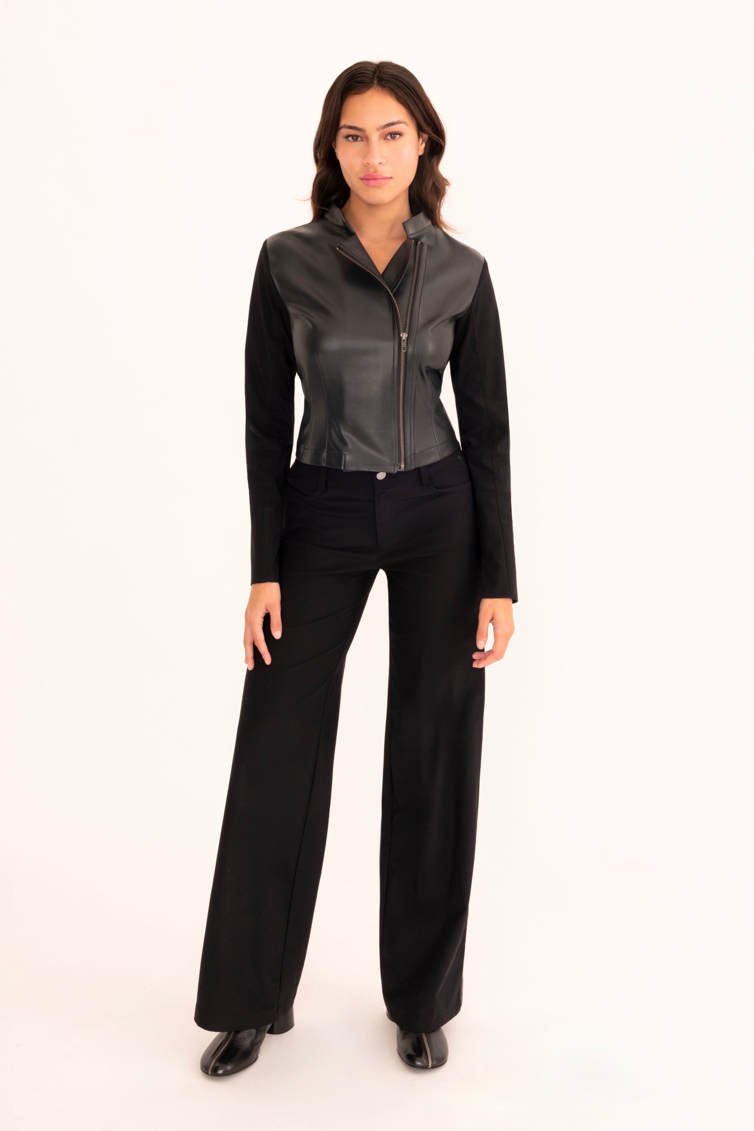 A woman modeling a black faux-leather jacket and wide-leg black trousers, standing against a solid white studio background.