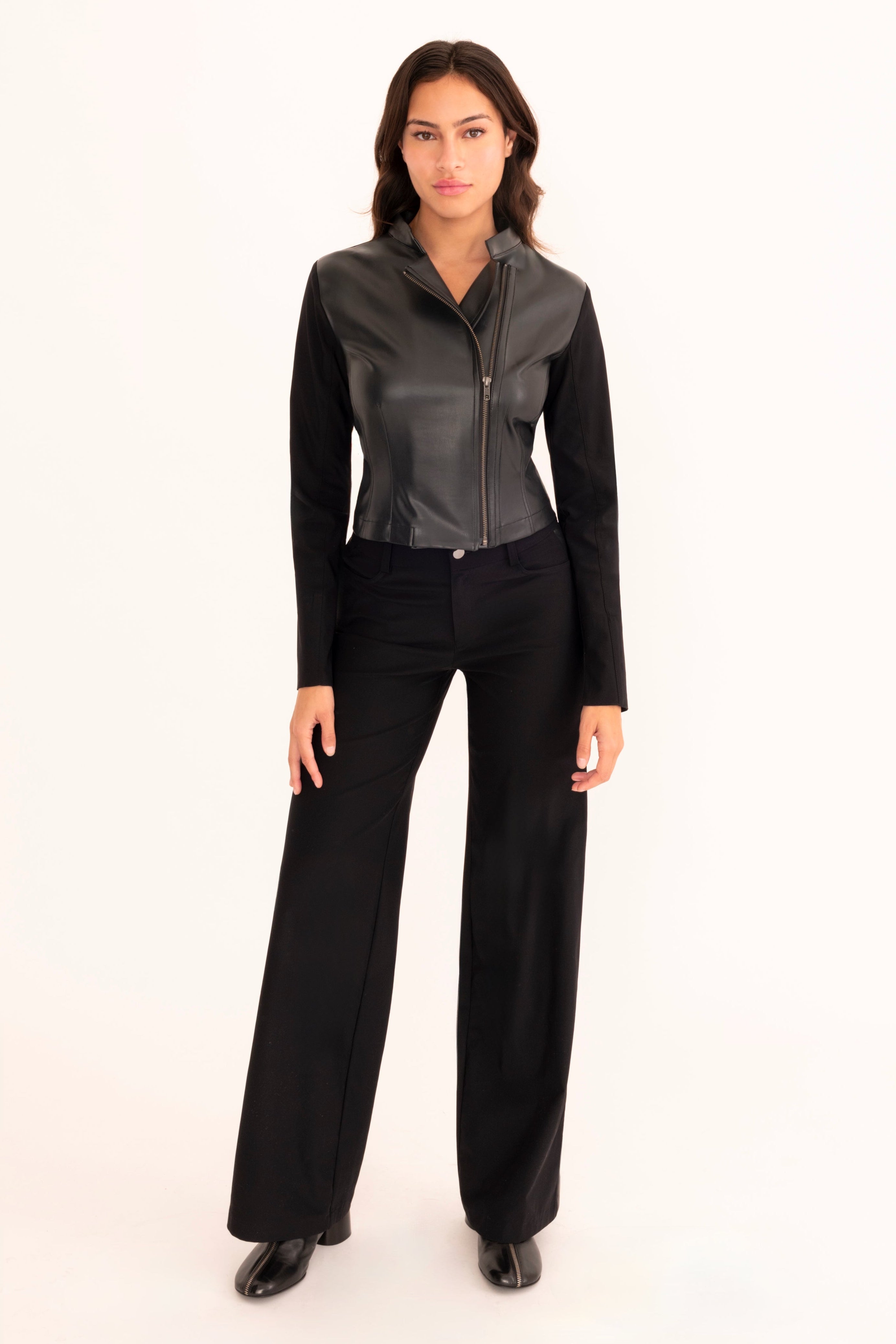 A woman modeling a black faux-leather motorcycle jacket with a stand collar and front zipper, paired with matching black pants, against a plain off-white studio background.