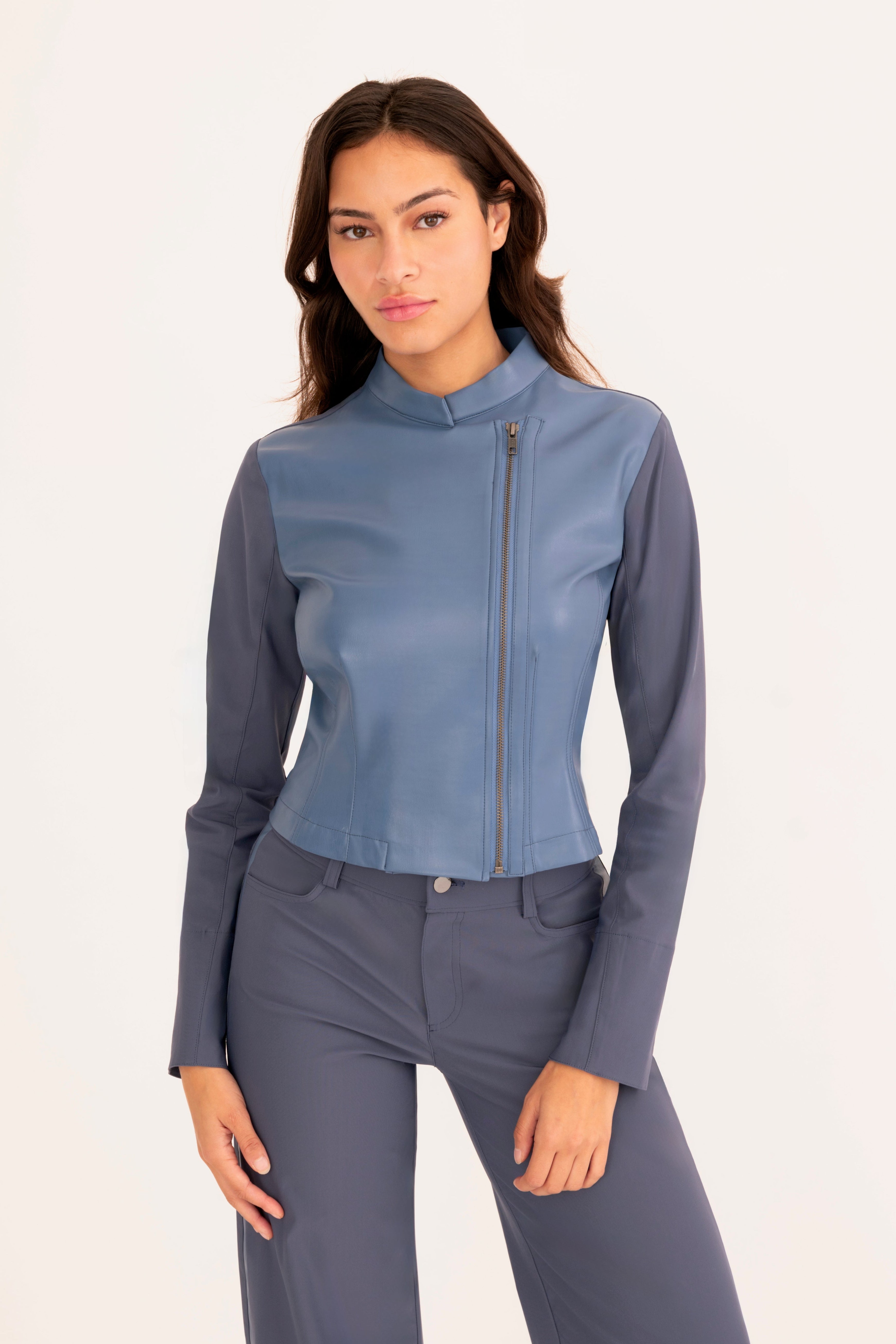 A woman modeling a blue faux-leather motorcycle jacket with a stand collar and front zipper, paired with matching blue pants, against a plain off-white studio background.