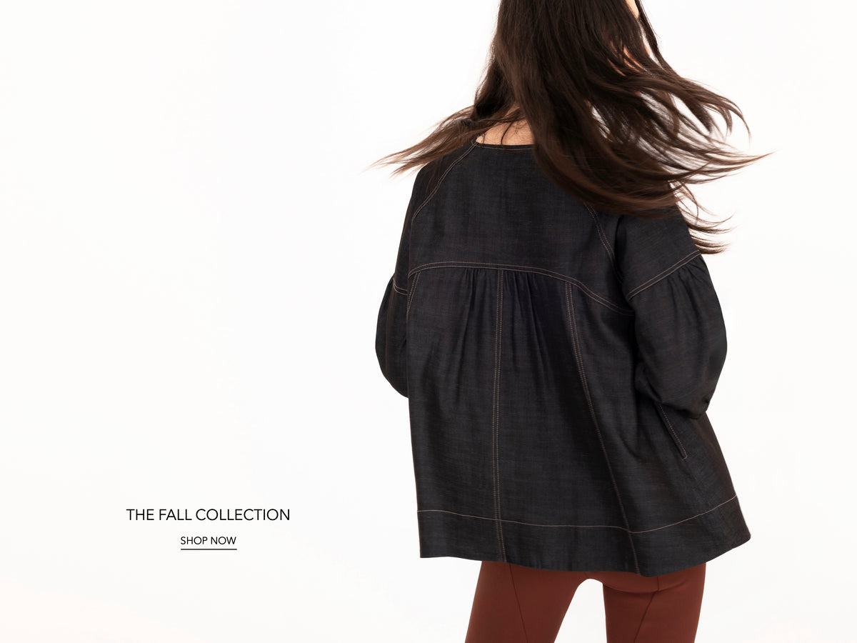 A woman shown from the back wearing a dark charcoal denim top with contrast stitching and brown pants, against a clean white background. Text on the left reads 'THE FALL COLLECTION' and 'SHOP NOW'.