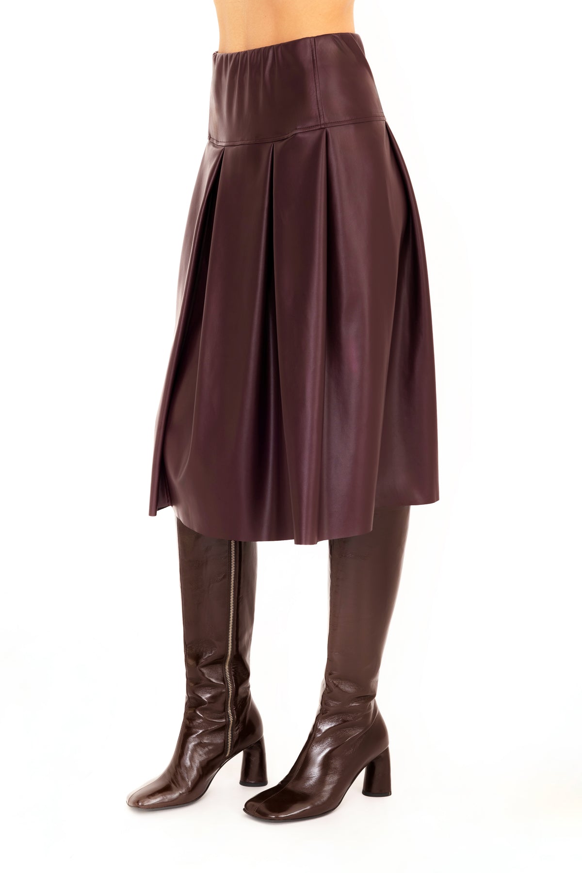 A burgundy vegan leather pleated A-line skirt shown on a model from the waist down, paired with dark brown knee-high leather boots against a plain white background.